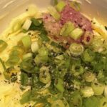 This image shows the ingredients for Garden Witch Radish Butter in the mixing stage. In a white bowl set on a wooden cutting board, we see a generous amount of softened butter as the creamy base and a pile of grated radish (light pinkish-purple in color), adding a vivid and earthy tone. Also visible are chopped green onions (scallions) sprinkled throughout for a fresh, mild onion flavor and a dusting of cracked black pepper, sea salt, and dried herbs (possibly thyme or parsley), adding seasoning and aromatic depth. The ingredients are not yet mixed together, showing a vibrant and rustic preparation step that feels both cozy and intentional—fitting the mystical, tavern-core aesthetic.
