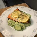 A white plate set on a wooden kitchen cutting board holds baked salmon served atop a creamy green salad drizzled with vinaigrette and garnished with herbs and lemon slices.