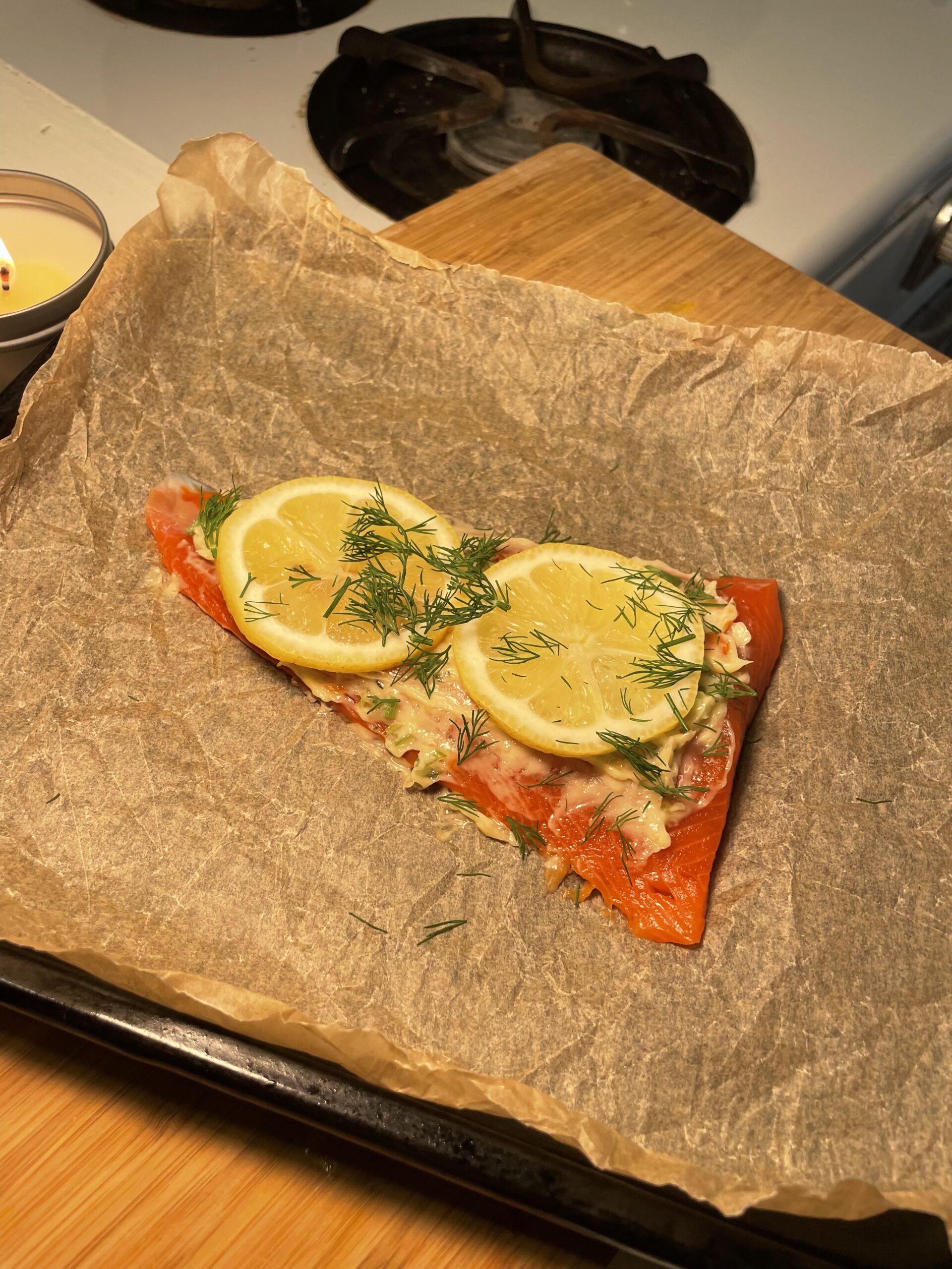 The salmon is placed on a sheet of brown parchment paper on a baking tray. It's topped with slices of fresh lemon and sprigs of fresh dill, with what appears to be a creamy or herb butter mixture spread over it. The scene is warmly lit, with a lit candle on the side and a wooden cutting board in the background, creating a cozy, inviting kitchen atmosphere. The stove burner in the background suggests it's about to go into the oven for roasting.