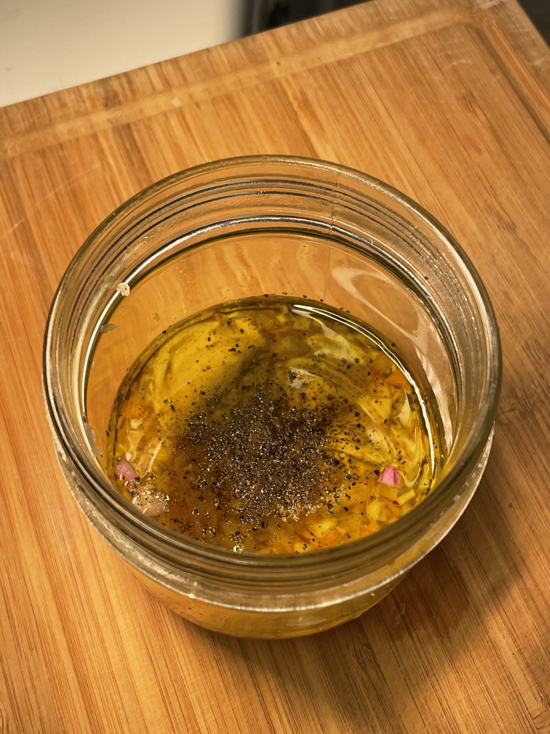 This image shows a small glass jar sitting on a wooden cutting board. Inside the jar is a golden-hued vinaigrette or marinade. It appears to be made of olive oil, finely chopped shallots or red onions, and cracked black pepper. The contents are unshaken, with the oil floating above the solids. This mixture looks like a fresh, homemade dressing—possibly a lemon-shallot vinaigrette—intended to accompany a salad and salmon dish.