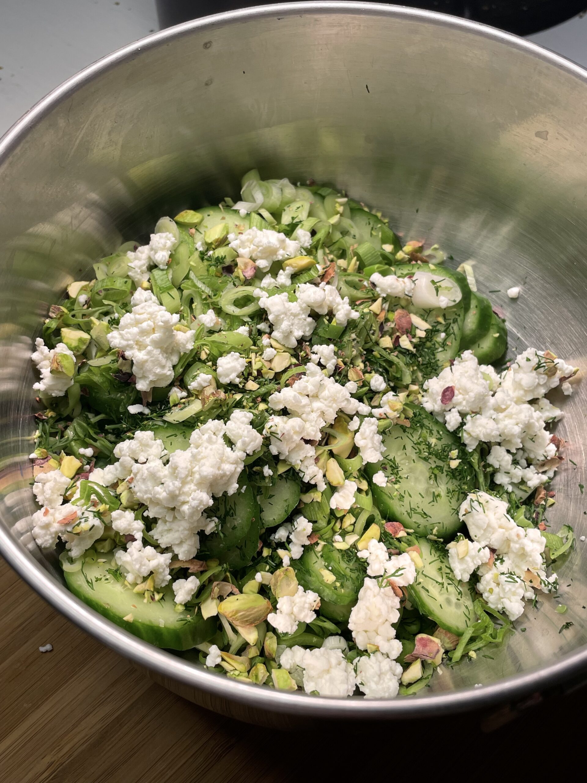 This image shows a fresh, vibrant salad in a large stainless steel mixing bowl. The salad features sliced cucumbers as the primary base, crumbled white cheese, chopped pistachios adding a crunchy, nutty element, chopped green onions (scallions), fresh chopped dill, contributing a bright, herby aroma.
The salad looks crisp and summery, with a blend of creamy, crunchy, and tangy components. 