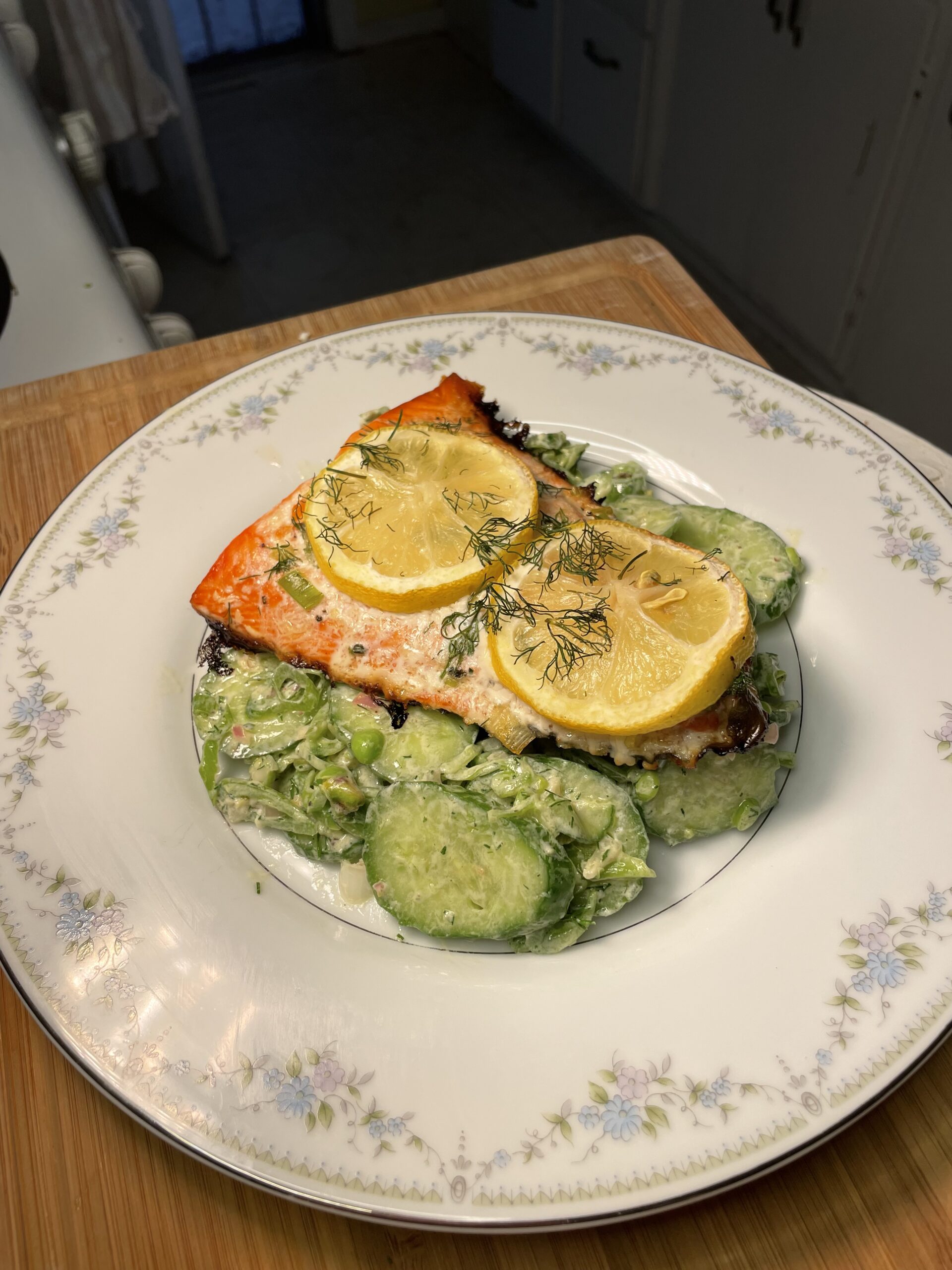 A white plate set on a wooden kitchen cutting board holds baked salmon served atop a creamy green salad drizzled with vinaigrette and garnished with herbs and lemon slices.