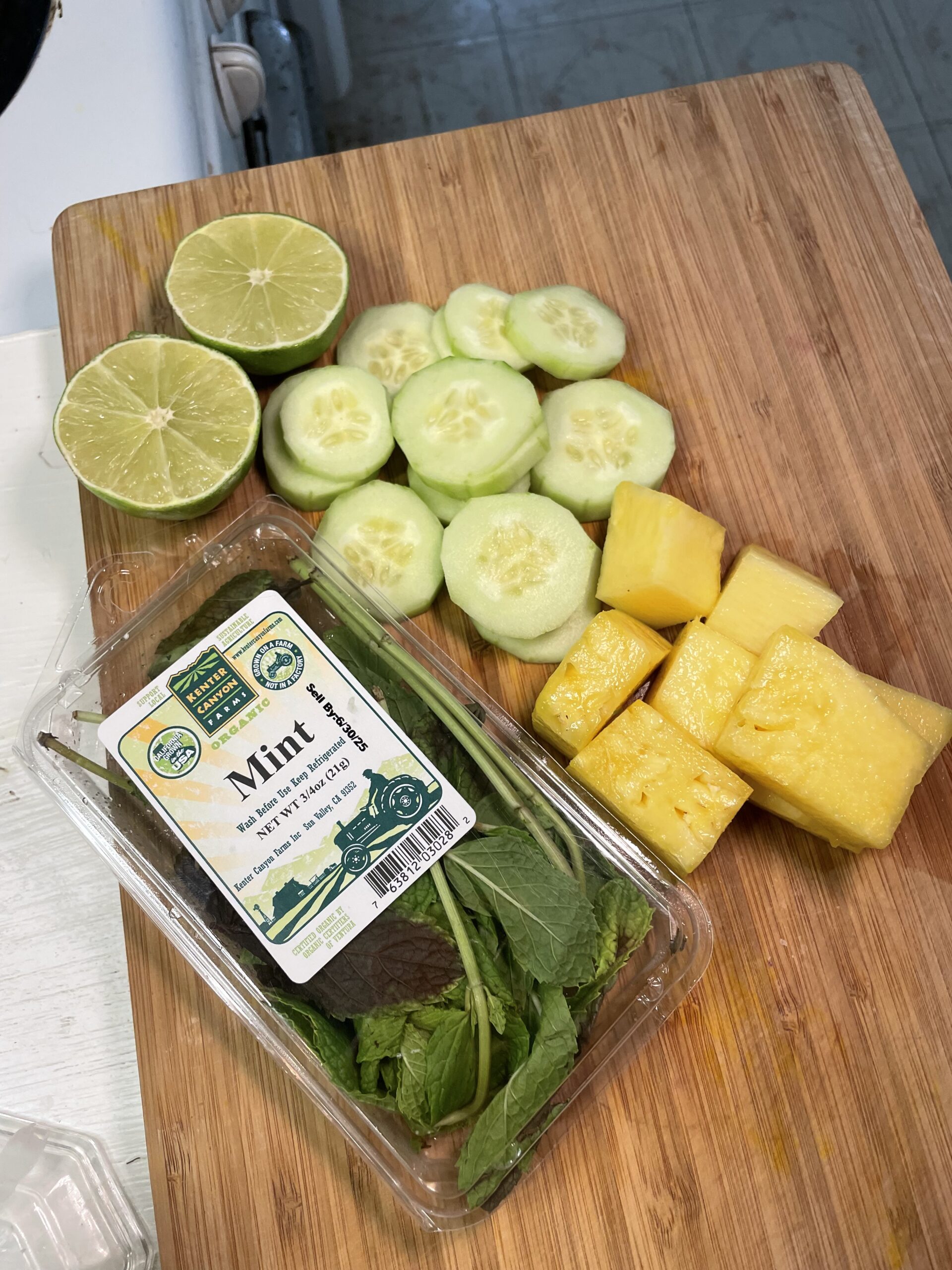 Pineapple chunks, cucumber slices, lime halves, and fresh mint in a plastic container, resting on a wooden cutting board in a kitchen setting. 
