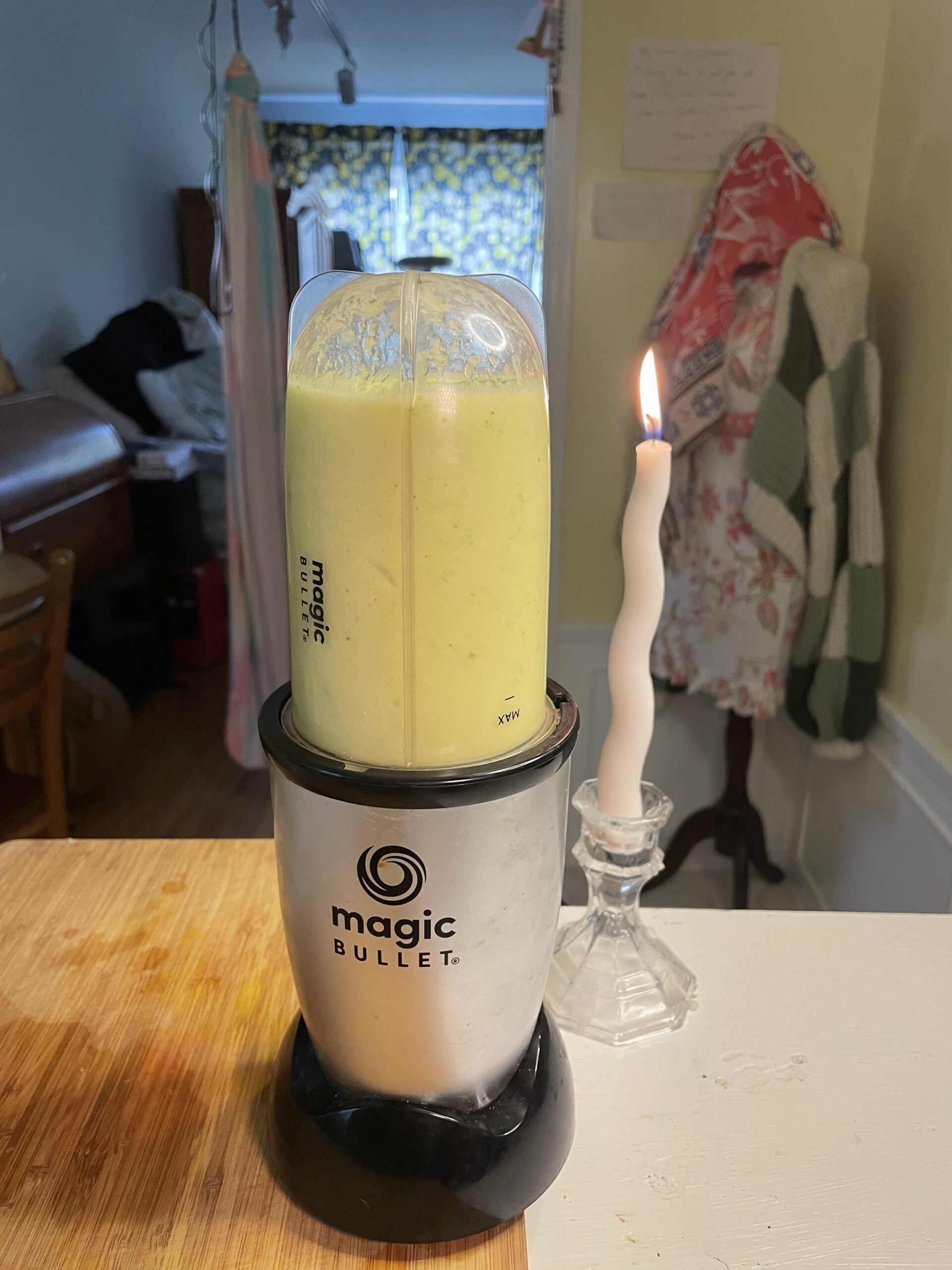 Green drink made of fresh pineapple chunks, cucumber slices, freshly squeezed lime juice, and fresh mint blended in a silver and black magic bullet blender. The blender is sitting on a wood cutting board next to a whimsical curved, spiral, white, lit candle in a beautifully decorated kitchen. 