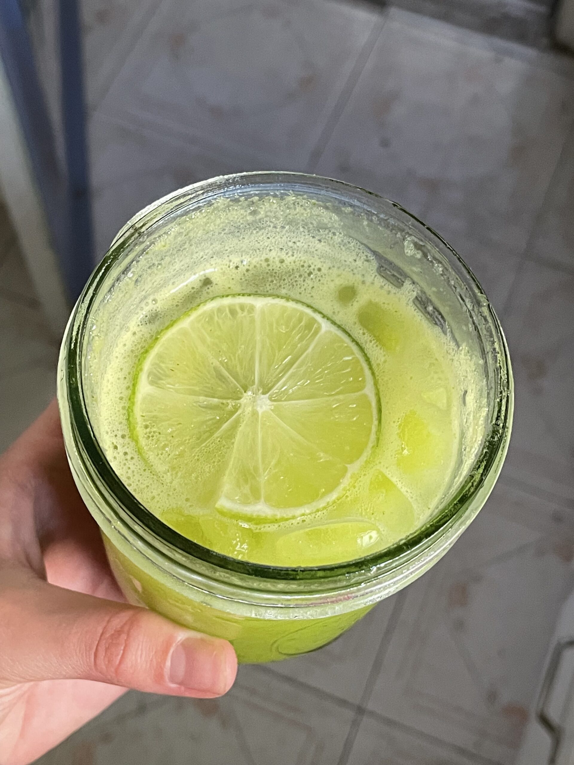 A fresh green drink in a glass cup is at the center. In the drink, there is a slice of fresh lime and ice. Because of the blending in the previous step, there is micro foam throughout the fresh green tonic. 