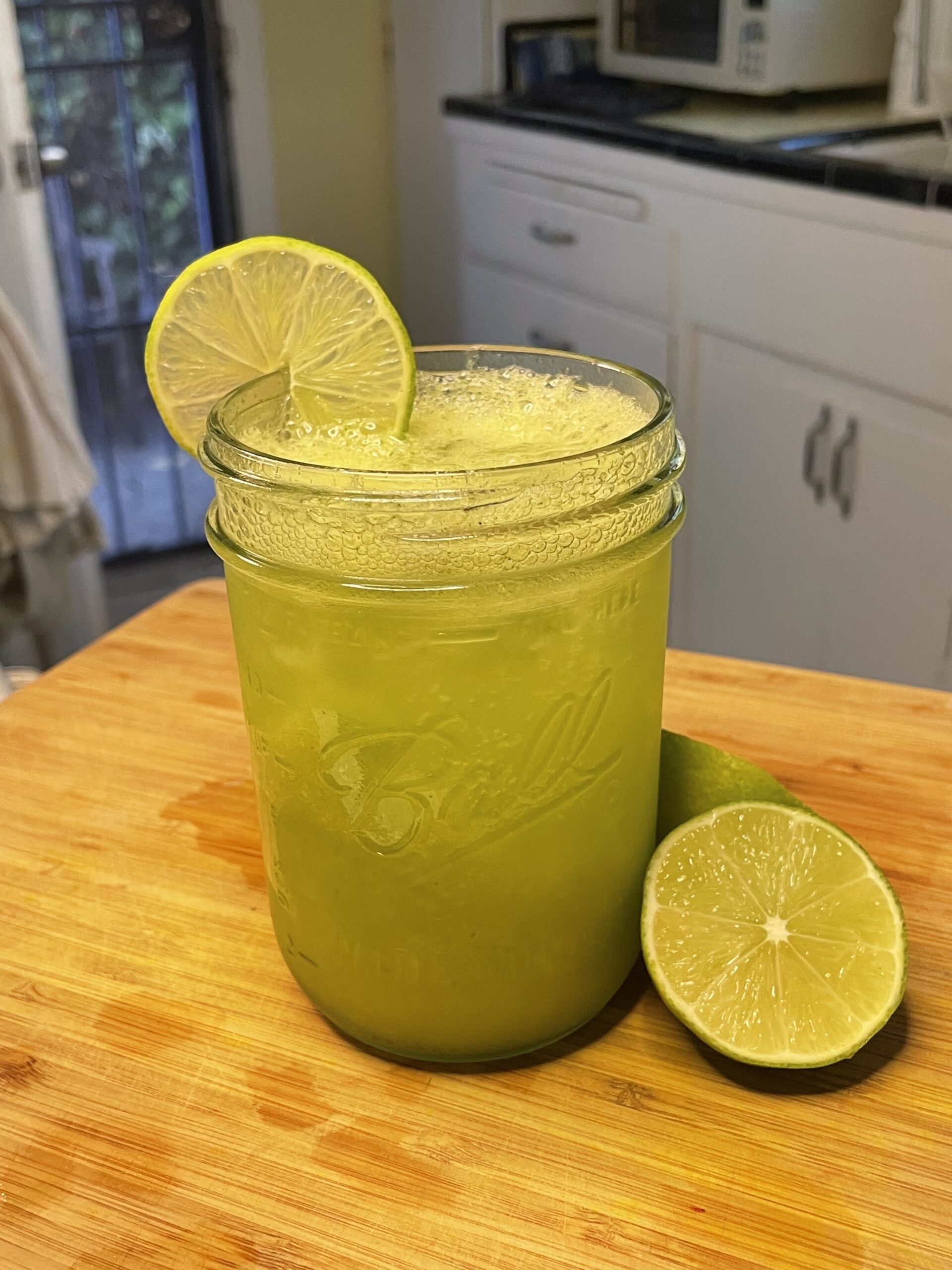 A refreshing, vibrant green sparking drink in a mason jar sitting on a wooden cutting board in a white kitchen. Sitting next to the hydrating drink is two juicy lime halves and a lime slice garnishing the drink. 