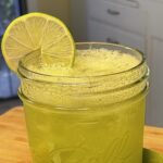 A refreshing, vibrant green sparking drink in a mason jar sitting on a wooden cutting board in a white kitchen. Sitting next to the hydrating drink is two juicy lime halves and a lime slice garnishing the drink.