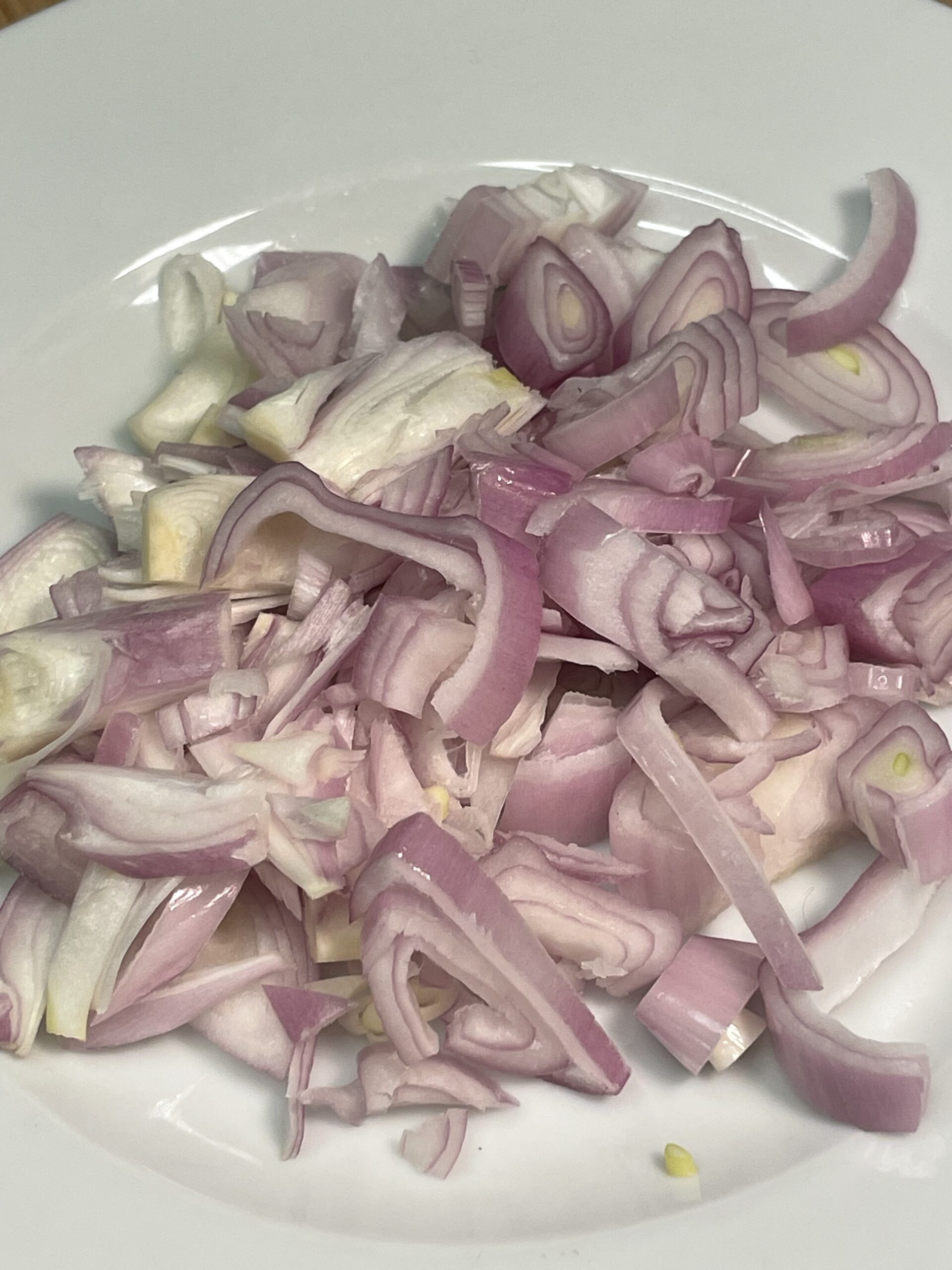 This image features a close-up of a plate filled with sliced shallots. The shallots are thinly and unevenly sliced, showcasing their beautiful pale purple and white layers. Their glossy surface suggests they’re fresh and ready for cooking—to be caramelized as part of the flavor base in a savory-sweet recipe.