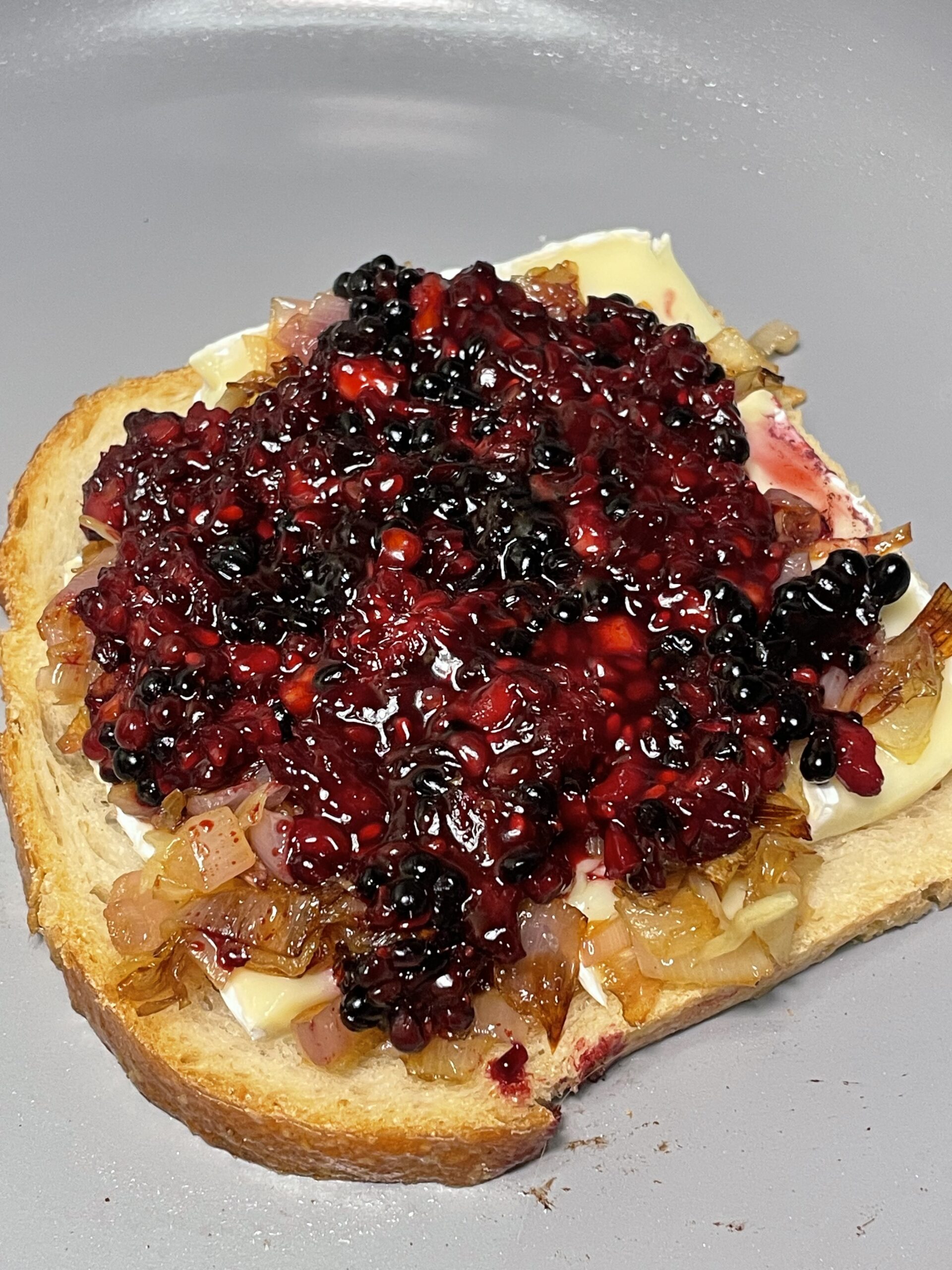 A piece of toast is covered in layers of a slice of brie, caramelized shallots, and blackberry jam compote rests in a non-stick pan.