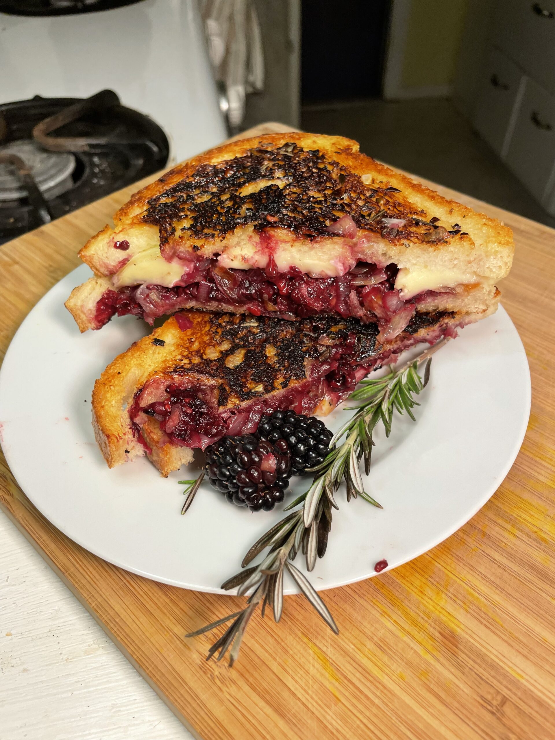 A beautifully styled Midnight Berry Brie Melt with Herbed Honey Butter rests on a white plate atop a wooden cutting board. The sandwich is grilled to a golden crisp (with some intentional or slightly charred edges), cut diagonally and stacked to reveal a decadent filling of melted brie cheese and a rich, dark berry compote—likely blackberries and shallots. A sprig of fresh rosemary and a whole blackberry are artfully placed in the foreground, enhancing the rustic and herbal aesthetic. The golden-buttered bread glistens, and the oozy, jammy berries lend a vibrant, indulgent contrast, making this dish visually striking and mouthwateringly rich.