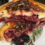 This image showcases a beautifully styled Midnight Berry Brie Melt with Herbed Honey Butter. The sandwich is grilled to a golden crisp (with some intentional or slightly charred edges), cut diagonally and stacked to reveal a decadent filling of melted brie cheese and a rich, dark berry compote—likely blackberries and possibly red onions or shallots, adding depth and texture. The sandwich is plated on a white dish set on a wooden cutting board. A sprig of fresh rosemary and a whole blackberry are artfully placed in the foreground, enhancing the rustic and herbal aesthetic. The golden-buttered bread glistens, and the oozy, jammy berries lend a vibrant, indulgent contrast, making this dish visually striking and mouthwateringly rich.