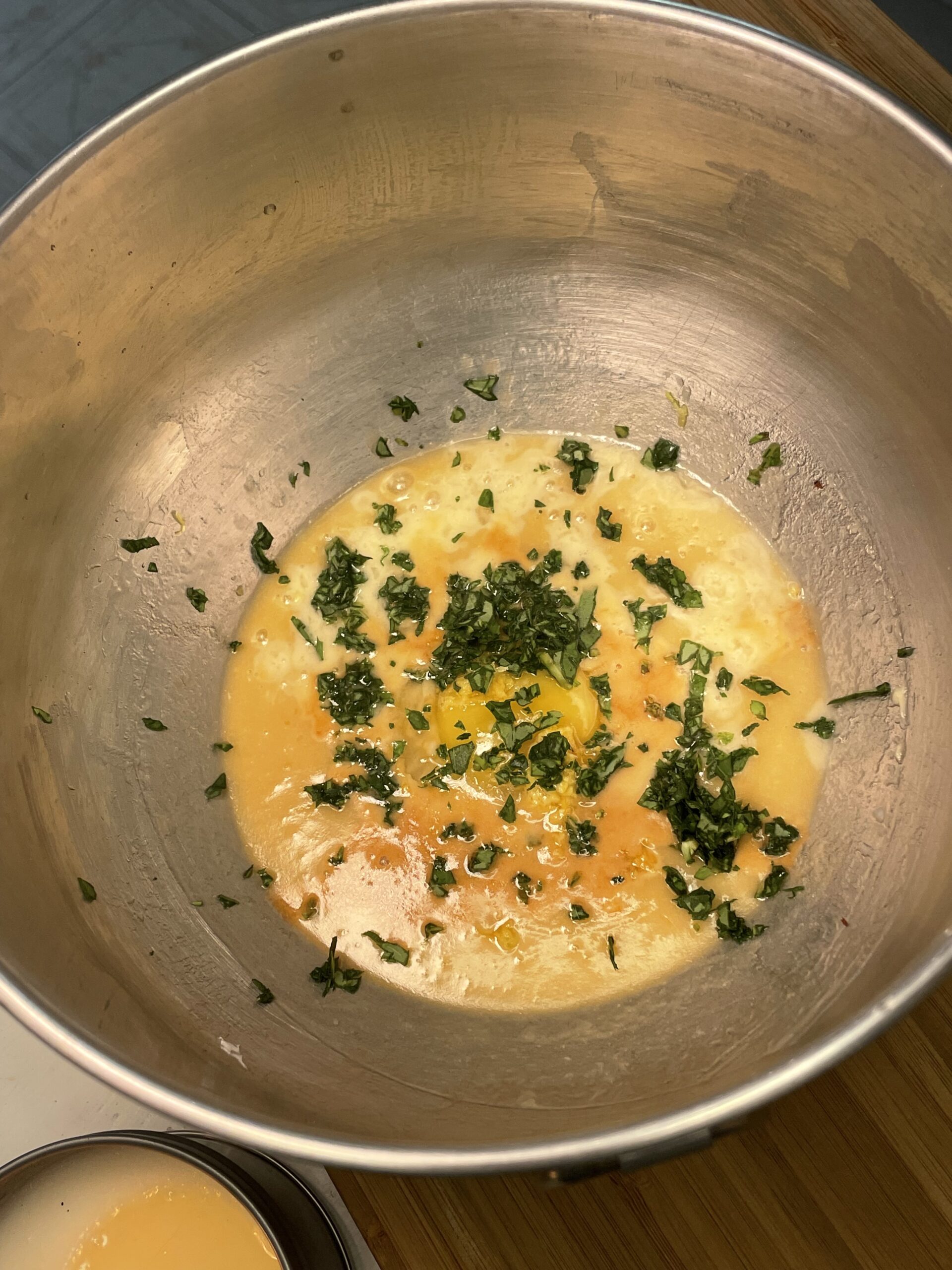 A metallic mixing bowl rests on a wooden kitchen countertop filled with the dough ingredients (eggs, lemon juice, basil, etc) to make Summer Basil Sugar Cookies.  This is the first step in the recipe's process.