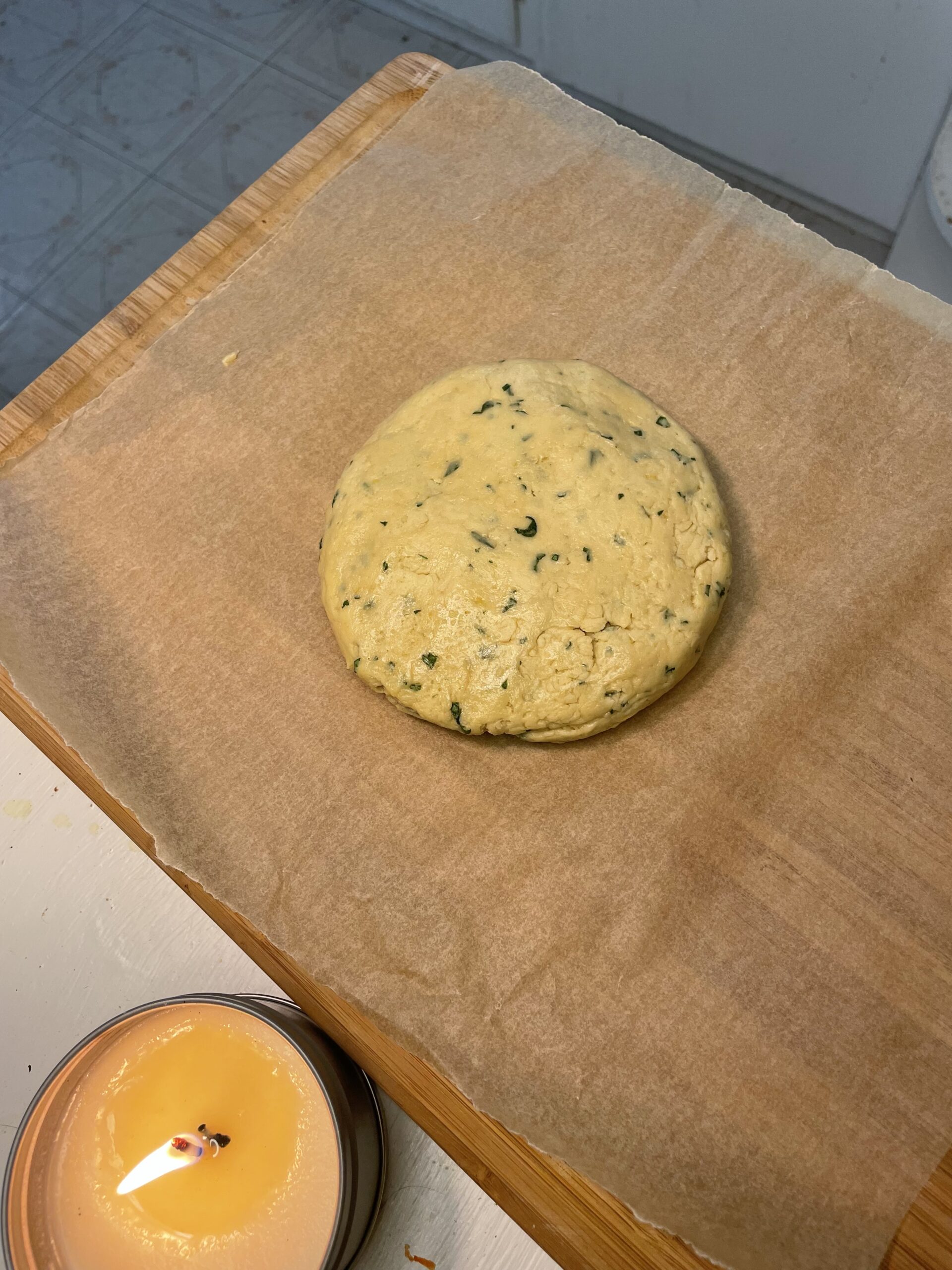 This image shows the prepared Summer Basil Sugar Cookies dough in a large disc formation, resting on a wooden cutting board covered with parchment paper.  The image shows the dough's consistency before being divided into individual cookies.  The dough is a soft yellow with specks of chopped basil throughout.  A candle rests on the kitchen counter in the corner for a homey feel.