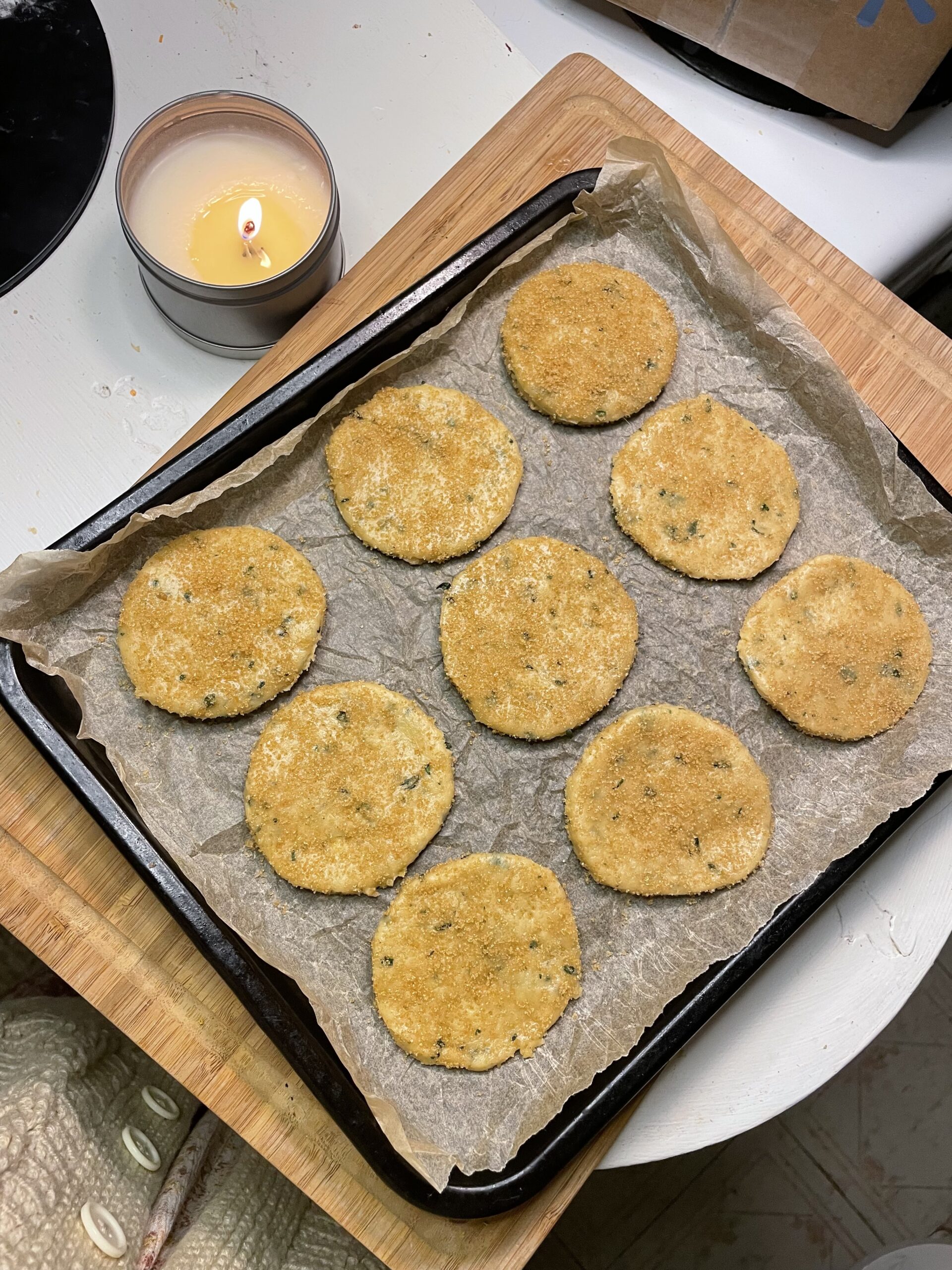 This image captures a cozy, pre-bake moment featuring a tray of unbaked Summer Basil Sugar Cookies arranged neatly on parchment paper. The cookies are evenly shaped into thick, round discs with a slightly textured surface, dusted with brown sugar. Small flecks of fresh basil are visible throughout the dough, hinting at the herbaceous flavor these cookies will offer.