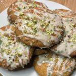 This image shows a plate of Summer Basil Sugar Cookies, beautifully presented on a wooden cutting board. The cookies are golden brown, with a slightly crisp exterior and a rustic, homemade shape. Each cookie is generously drizzled with a creamy white glaze and topped with a scattering of chopped pistachios, adding texture, color, and a hint of elegance.