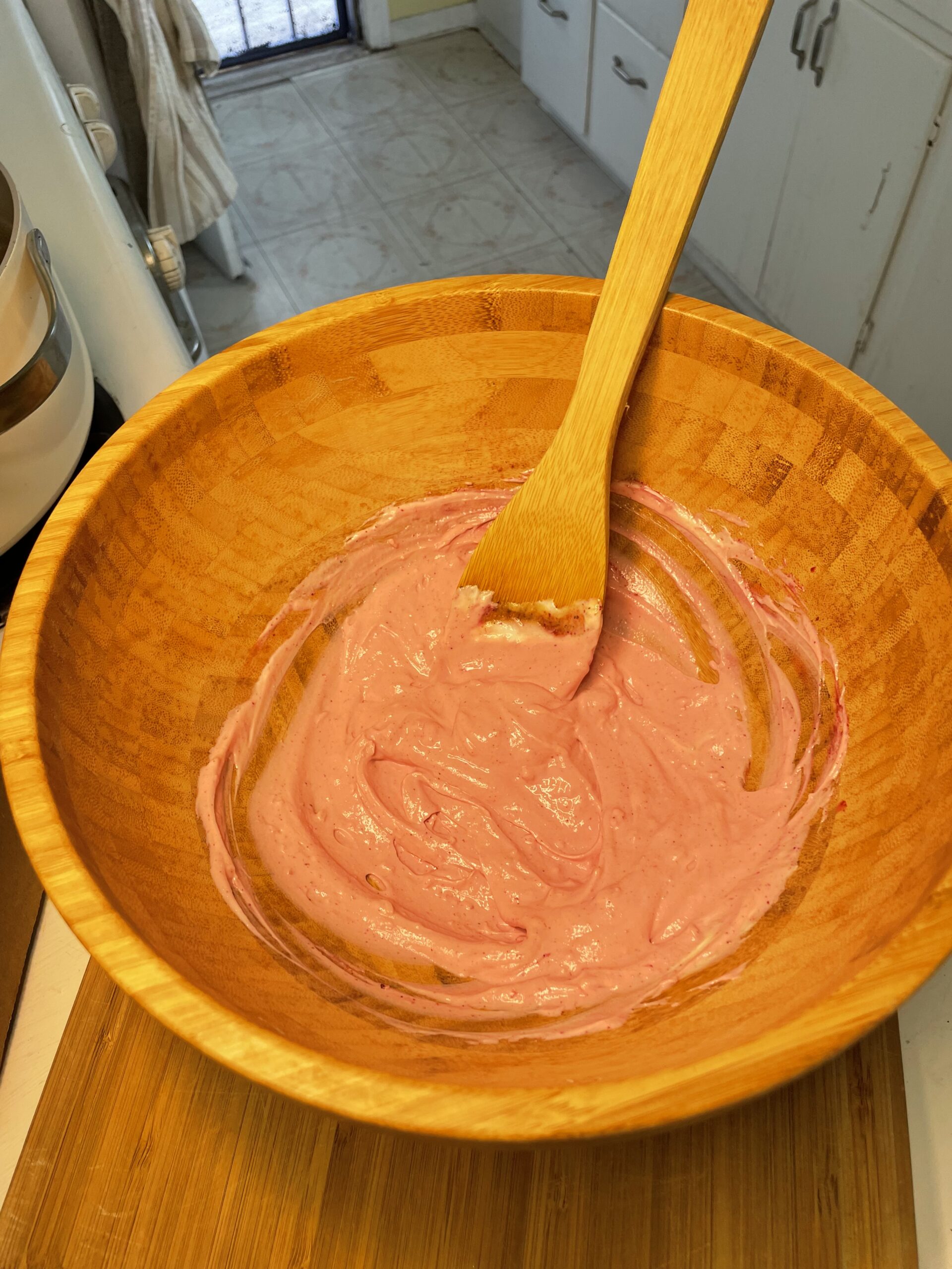 A large wooden bowl holds a creamy pink dressing mixture being stirred with a wooden spatula. The vibrant hue suggests the presence of beet juice or another naturally colorful ingredient, likely used to give the salad its signature "blush" tone.