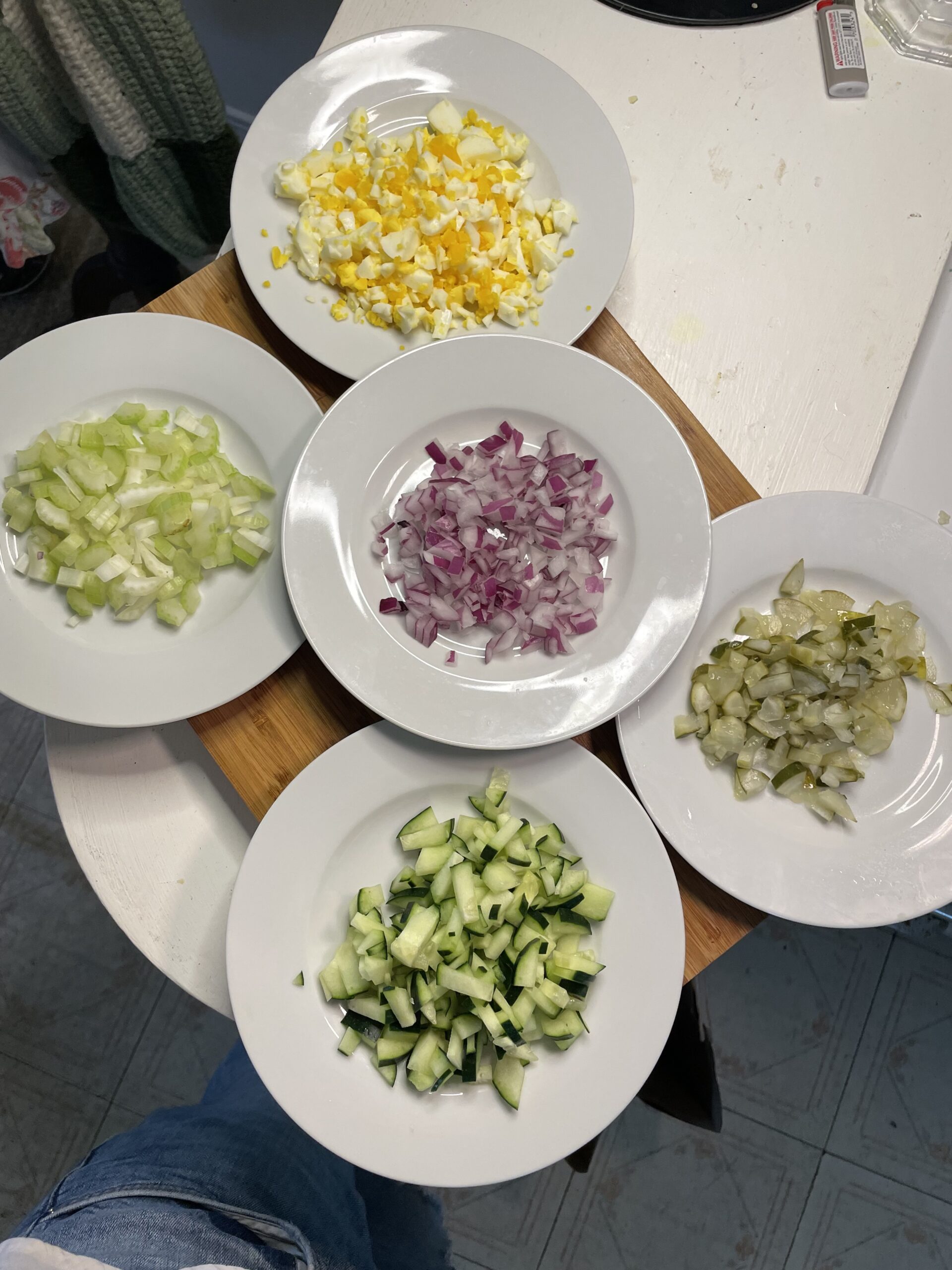 Five small white plates are neatly arranged on a counter, each showcasing a different chopped ingredient, diced celery (top left), chopped hard-boiled eggs (top right), chopped pickles (bottom right), chopped red onion (center), and diced cucumber (bottom). These mise en place-style shots highlight the fresh, crunchy elements that will soon be folded into the pink potato base—offering texture and flavor contrast.
