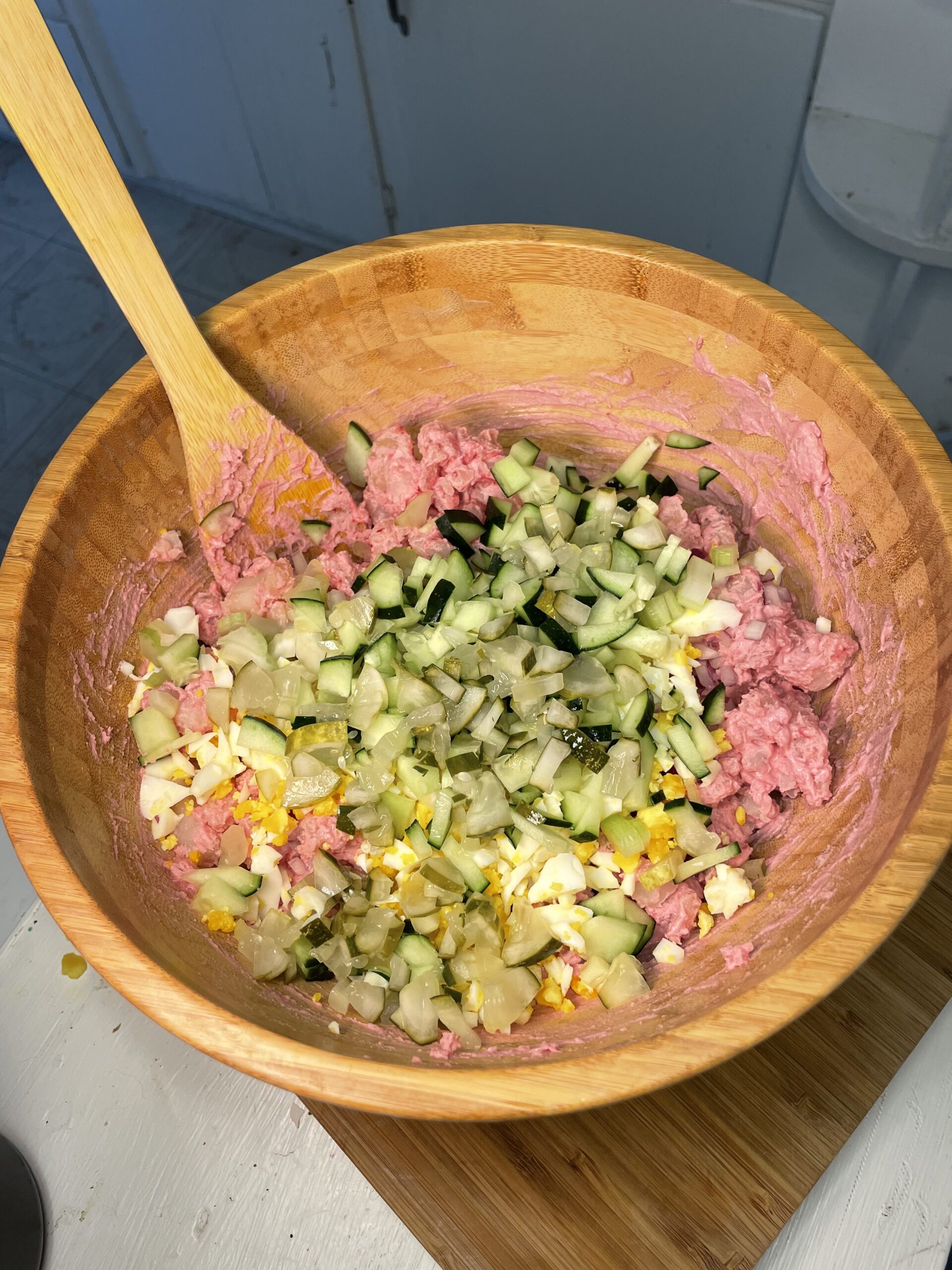 The same wooden bowl is now partially filled with the pink dressing and topped with an assortment of salad ingredients, including diced cucumber, chopped pickles, and crumbled hard-boiled eggs. Bits of the pink-dressed potatoes are visible underneath, ready to be folded together for a refreshing summer side dish.