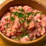 Redish pink potatoes sit atop a blend of chopped vegetables. This is the final product of the Summer blush potato salad. The salad, garnished with parsley, sits in brown wooden serving bowl, resting atop a wooden kitchen cutting board.