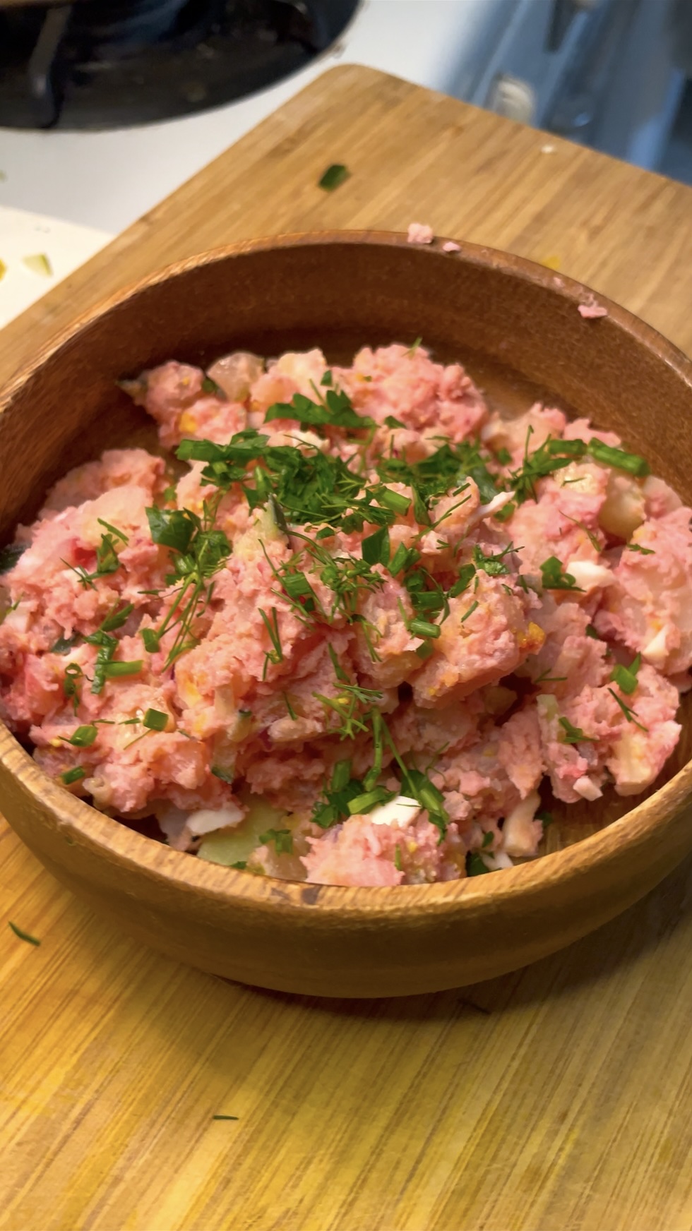 Redish pink potatoes sit atop a blend of chopped vegetables. This is the final product of the Summer blush potato salad. The salad, garnished with parsley, sits in brown wooden serving bowl, resting atop a wooden kitchen cutting board.