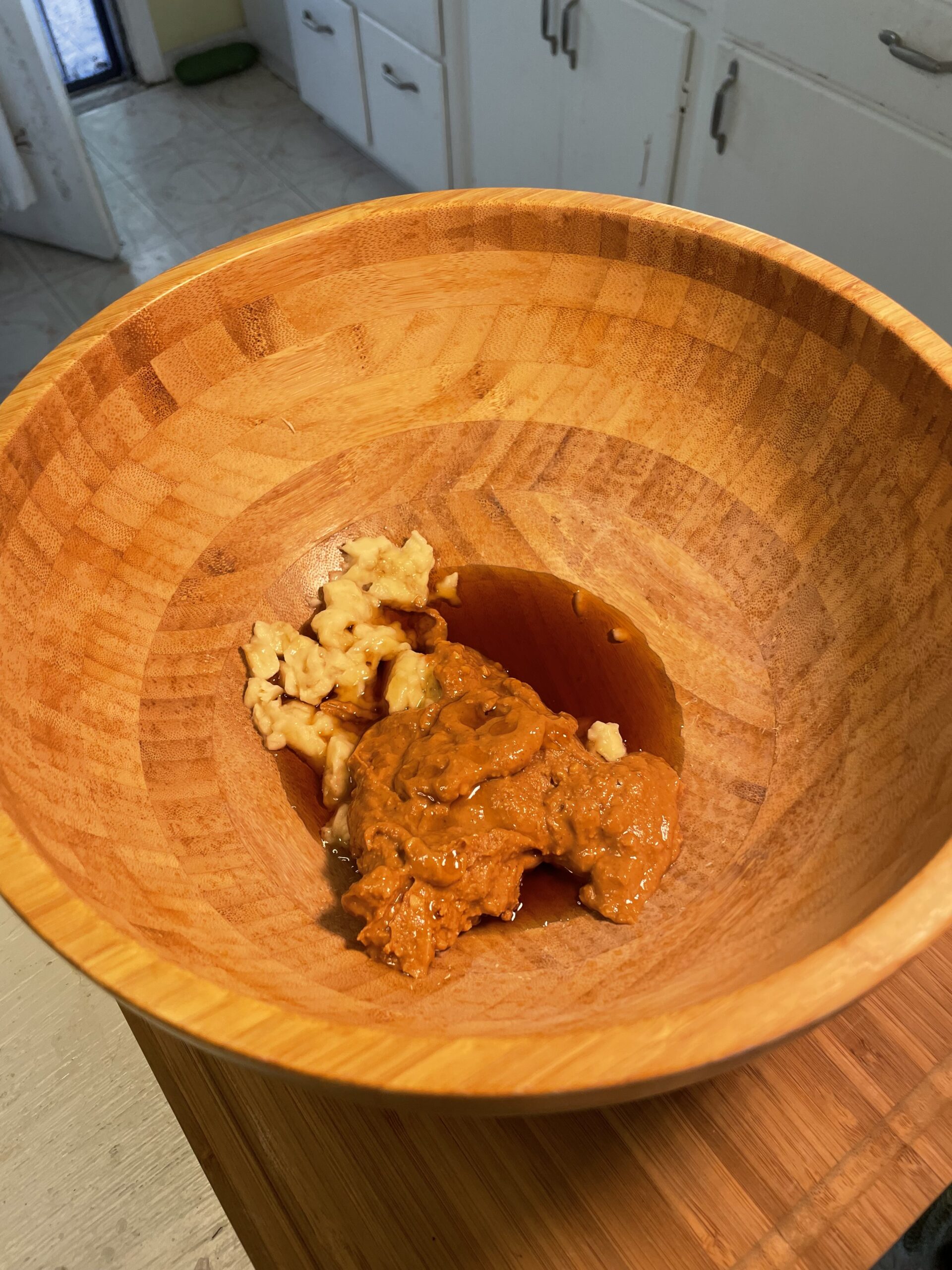 Large wooden mixing bowl filled with wet ingredients including almond butter, mashed banana, applesauce, maple syrup, honey, vanilla, coconut cream, oil, and nut milk.