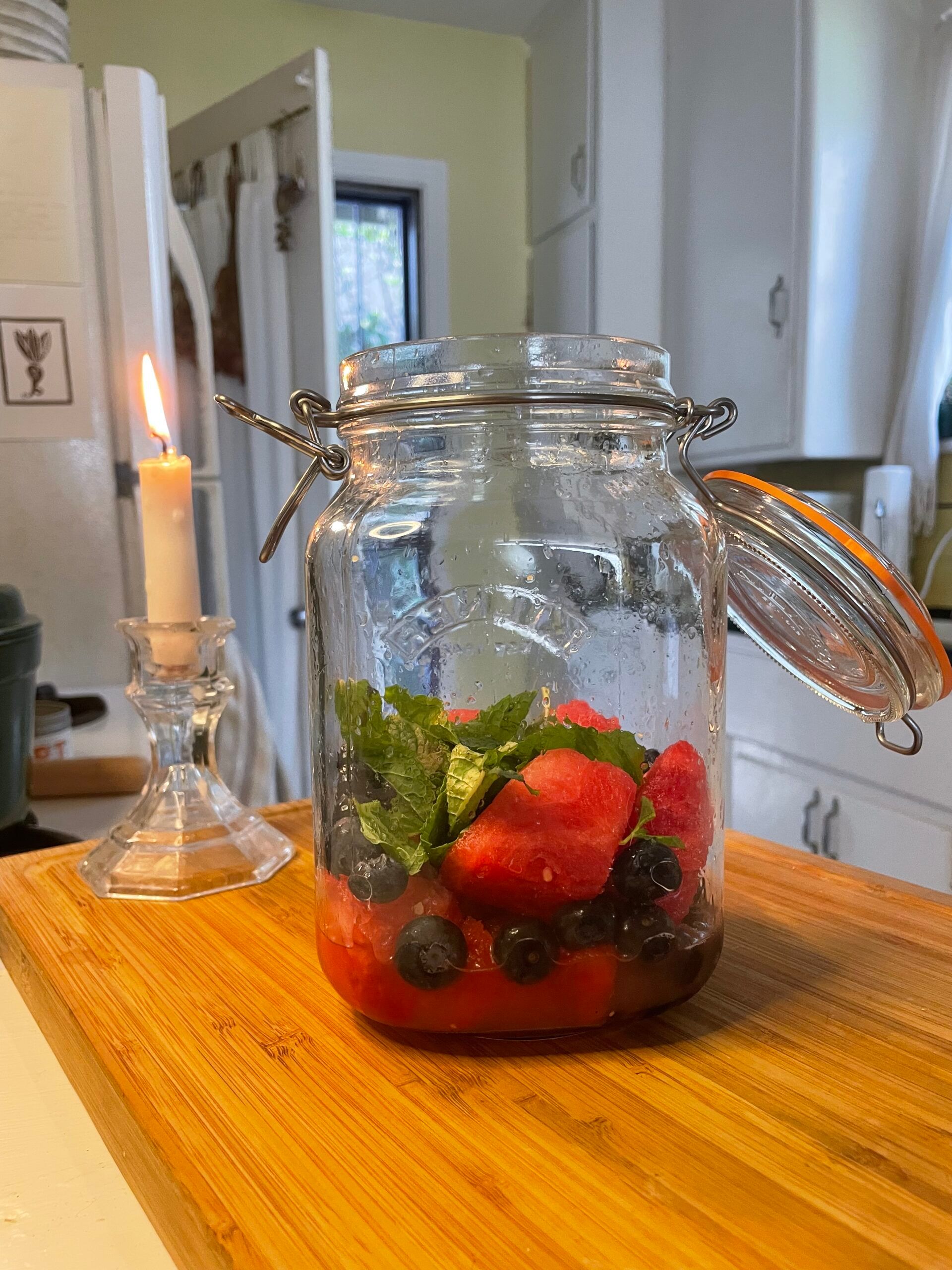 In a light kitchen, sitting on a wood cutting board, a large mason jar is halfway filled with summer fruits and mint: cubed watermelon, freshly squeezed lemon juice, and blueberries. Next to the jar is a whimsical, lit candle in a vintage glass candle holder. 