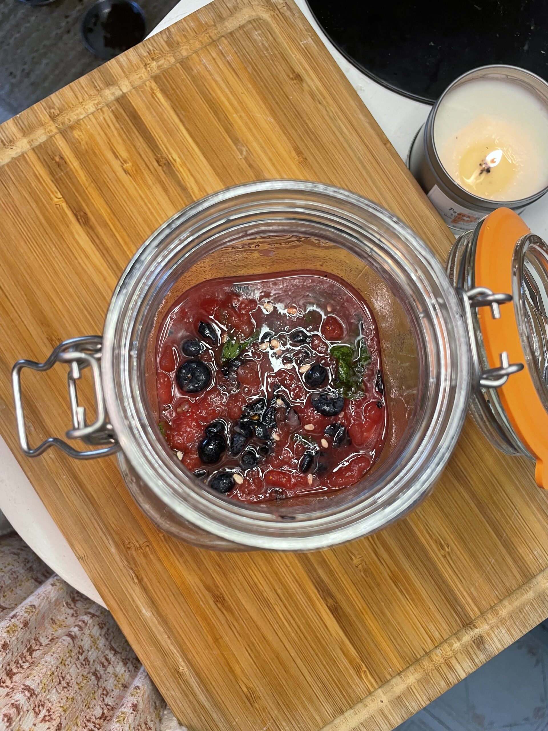 On a wooden cutting board, you can see the muddled fruits and herbs: freshly squeezed lemon juice, watermelon, blueberries, and mint from a top down view. Another lit candle is in view. 