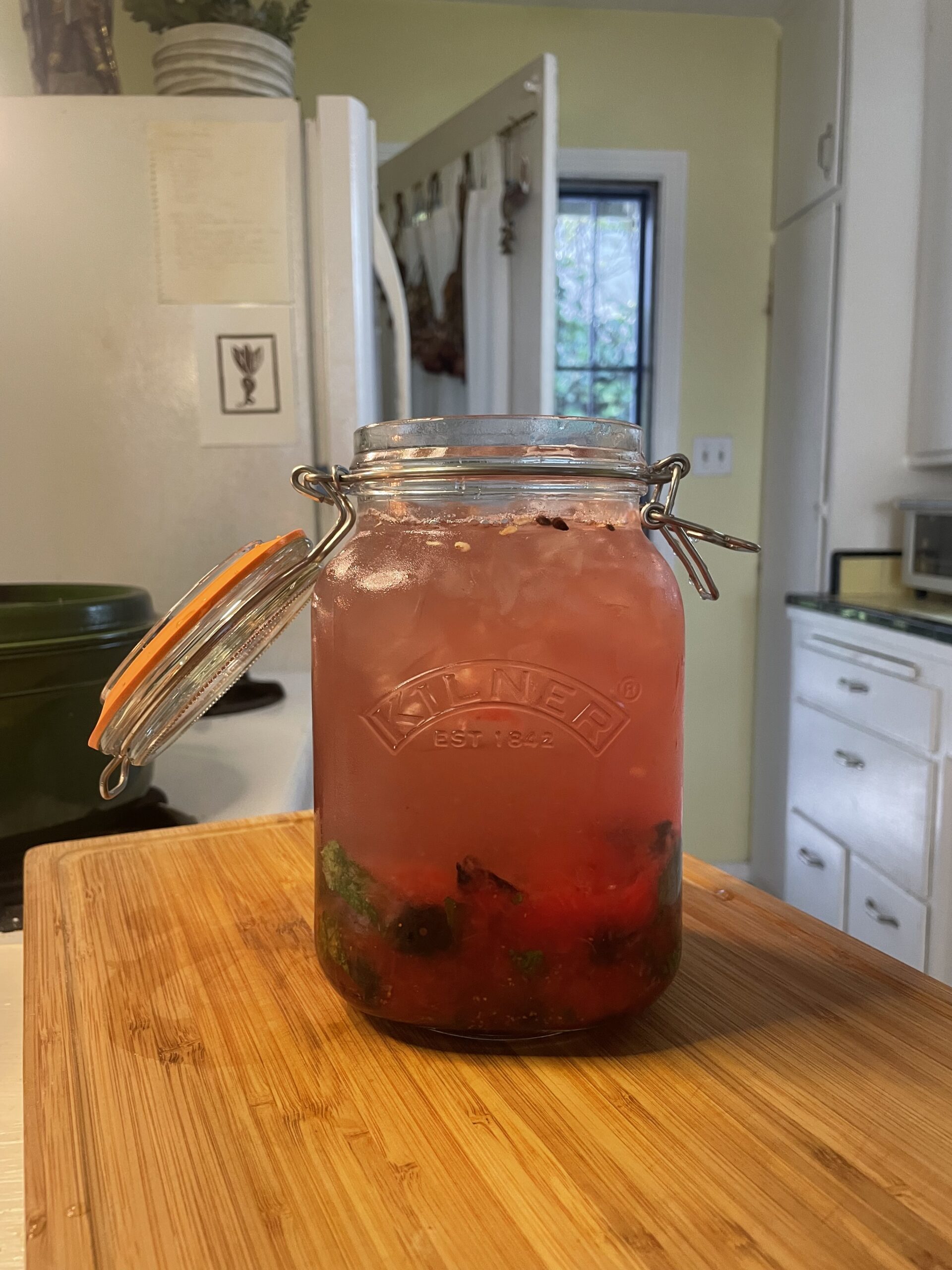 In a light, yellow and white kitchen, a large mason jar is sitting on top of wooden cutting board. The large mason jar is filled to the brim with muddled summer fruit including watermelon, blueberries, mint and lemon, with added hibiscus tea and coconut, stirred or shaken together with ice to create a refreshing drink. 