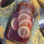 On an adventure, the fairy berry punch is a light pink, purple color with several yellow lemon slices visible in the jar. Around the punch are red cowboy boots, a coffee drink, a speaker, a yellow book, as well as a brightly colored yellow, blue, and purple flower blanket.