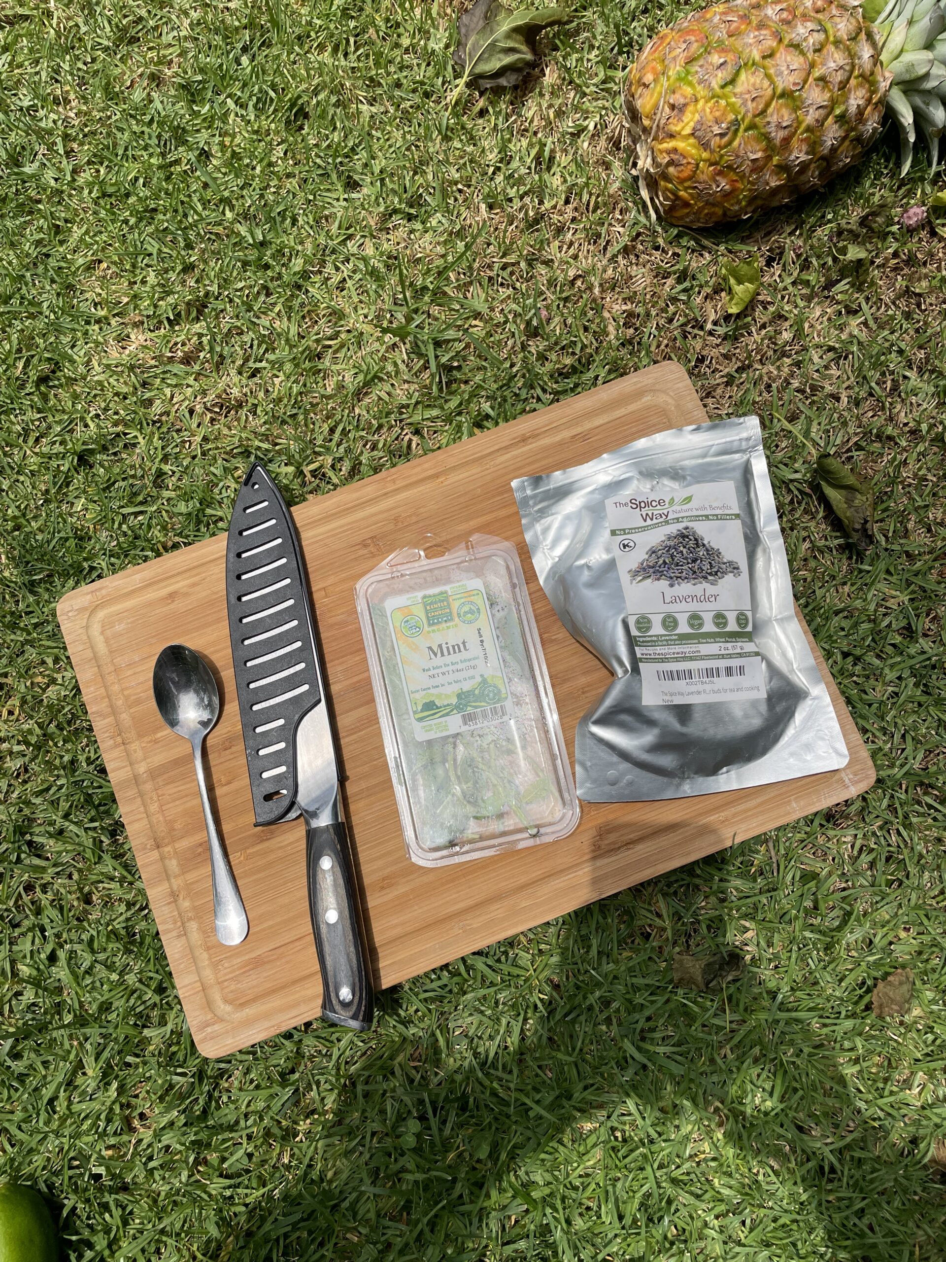 In a green field, a juicy pineapple is laying on the right side and a wooden cutting board is in the middle. On the cutting board is a metal spoon, a metal knife, a plastic package of mint and a metallic packet of lavender. 