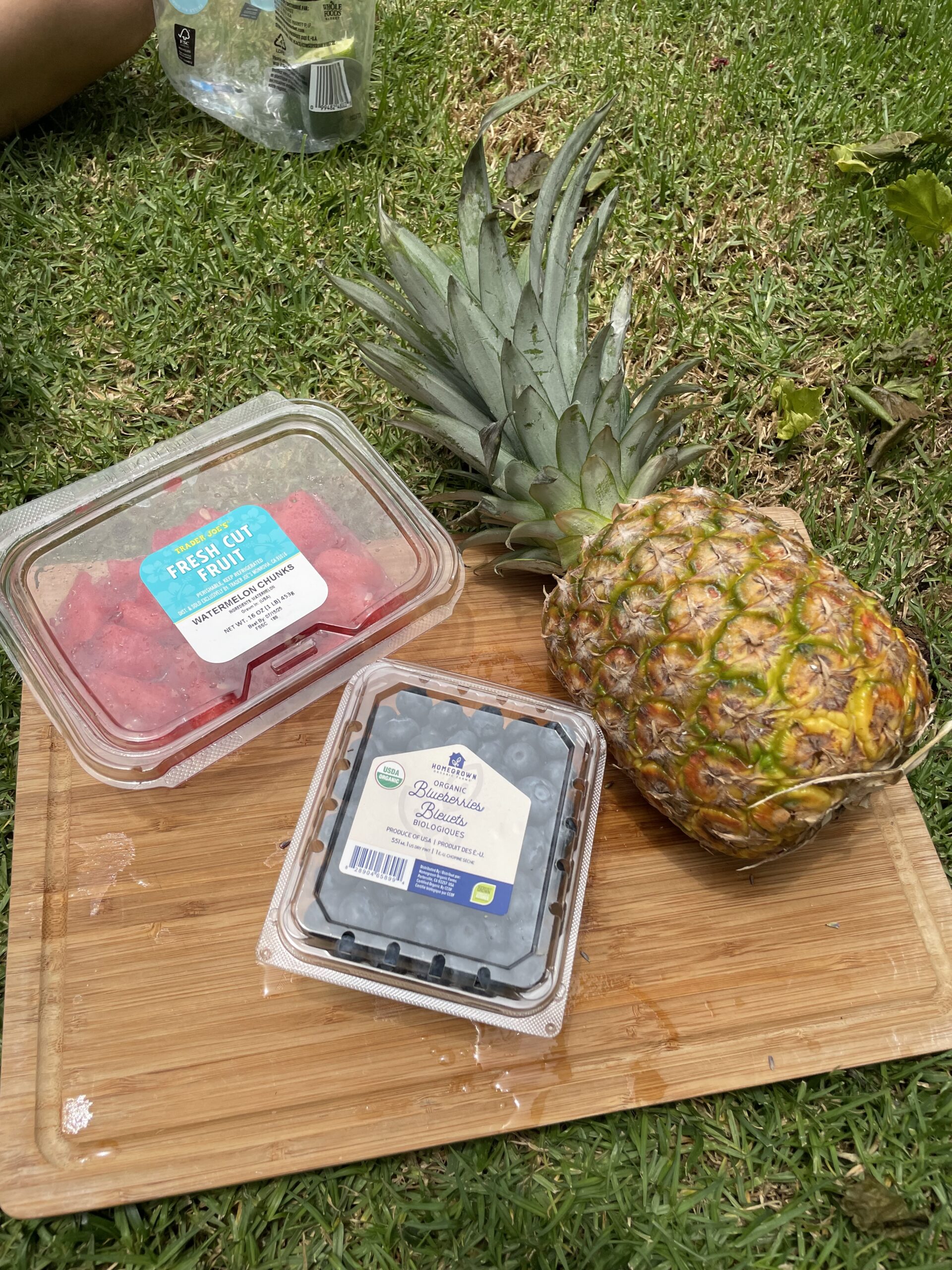 In a green field, a wooden cutting board has a large pineapple sitting on it as well as packaged cubed watermelon and blueberries. 