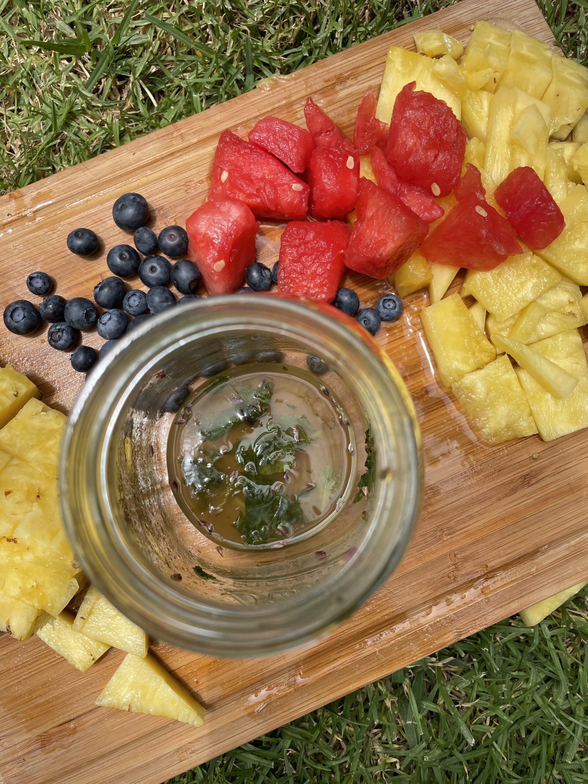In a green field, there is a wooden cutting board with cubed pineapple and watermelon, as well as blueberries and a glass jar. 