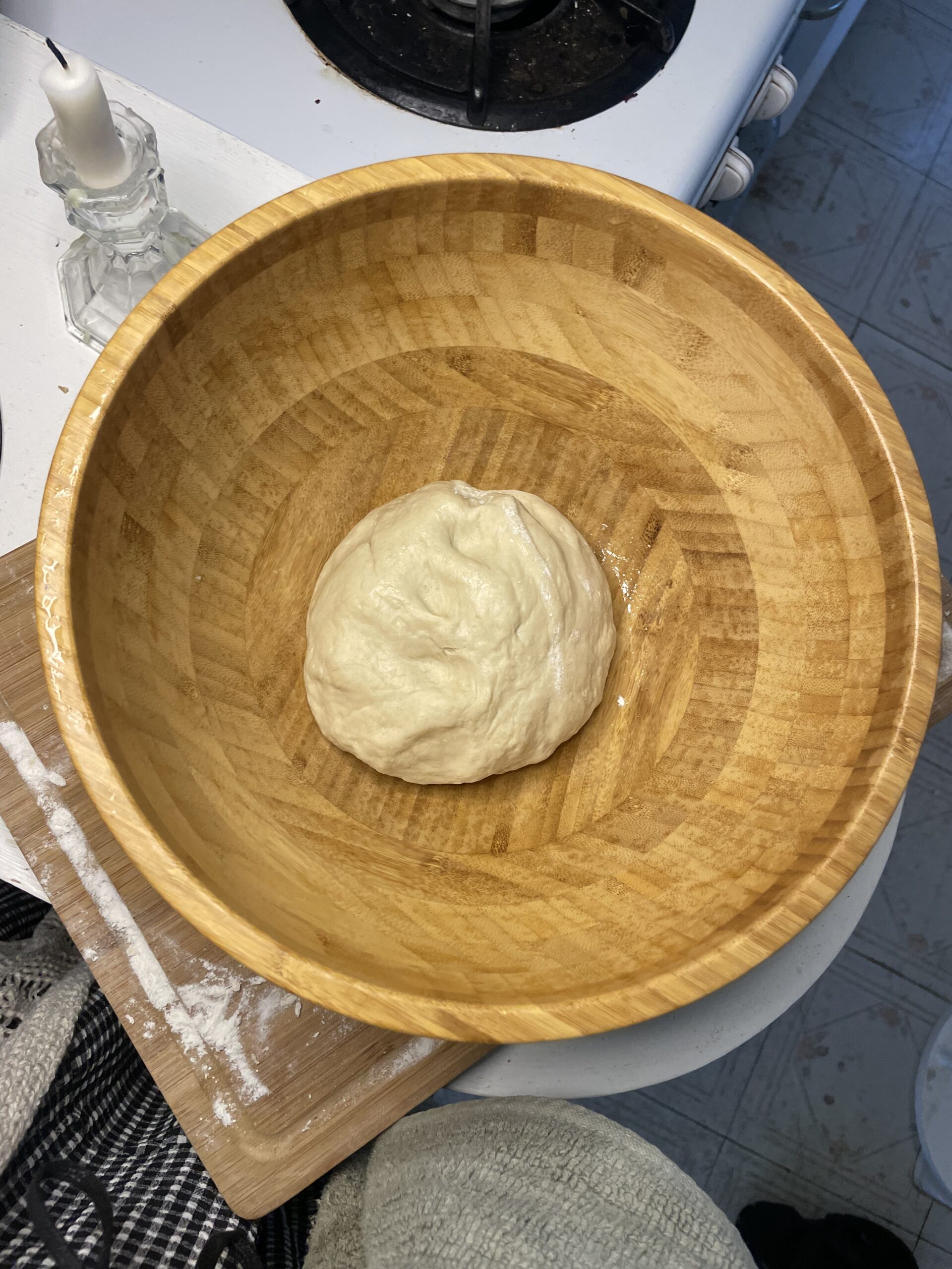 A fluffy ball of fresh, beige dough in the middle of a large, light wooden bowl sitting in a white kitchen next to a white candle in a glass candle holder. 