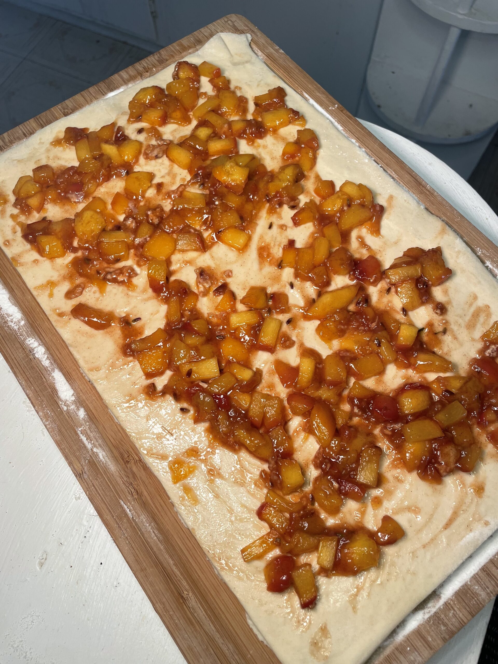 On fluffy dough lies the cut peach mixture full of sticky, sugary syrup on a wooden cutting board with white flour dusted on top. 