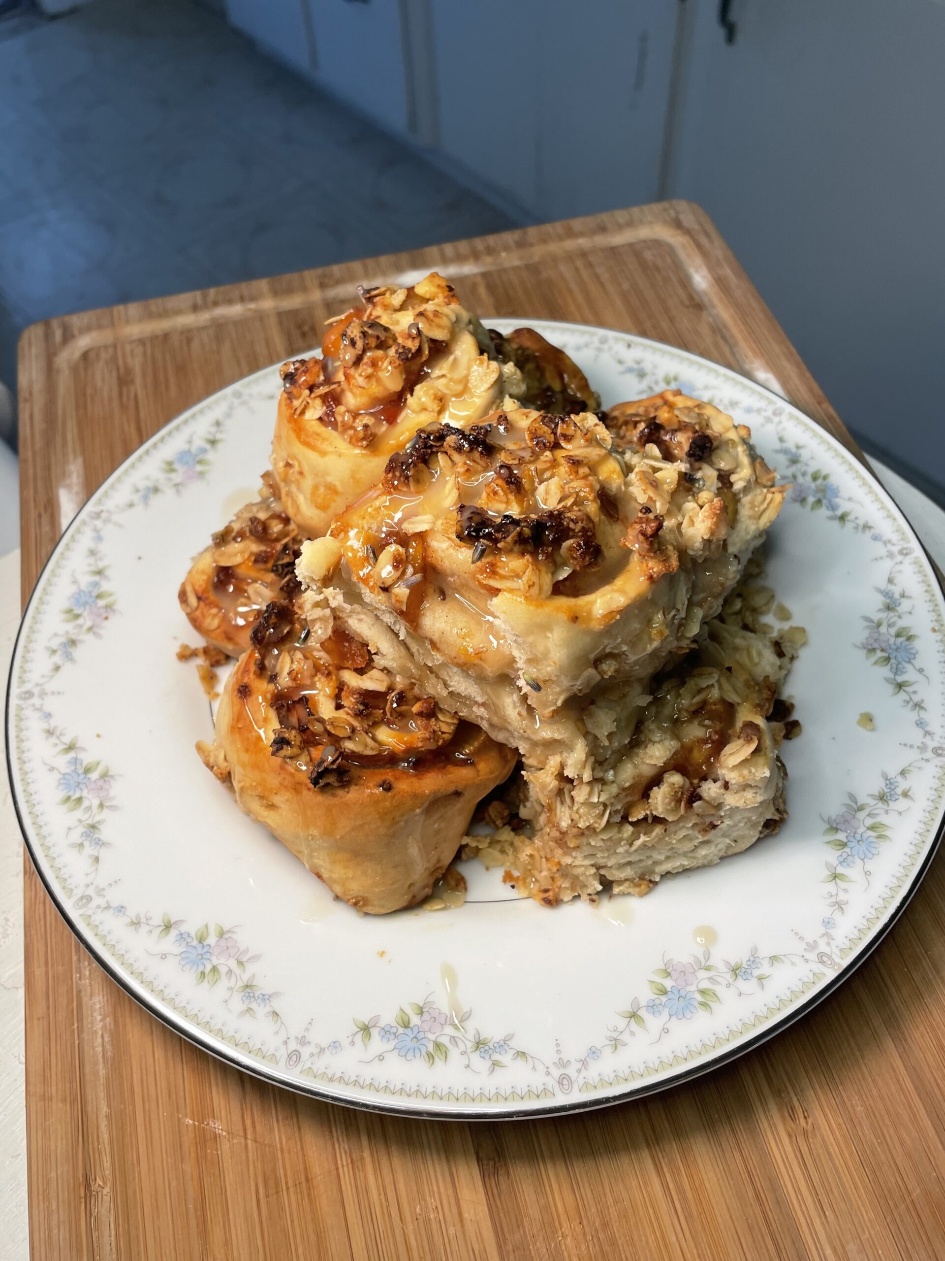 A pile of golden brown peach cinnamon rolls crusted with oats and coated sugary syrup sit on a white, floral plate. 