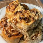 A pile of golden brown peach cinnamon rolls crusted with oats and coated sugary syrup sit on a white, floral plate.