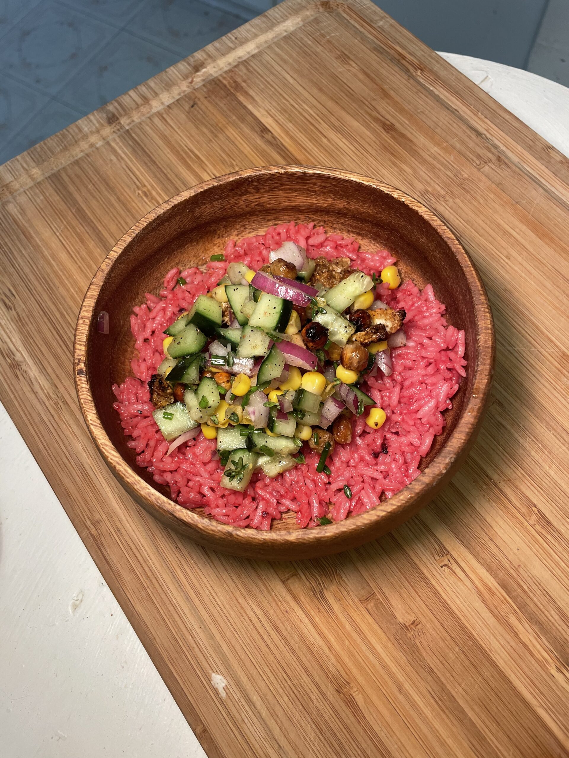 In a white kitchen, there is a wooden cutting board with a wooden bowl with pink rice and cooked green vegetables, chickpeas, corn, red onion, and herbs on top.