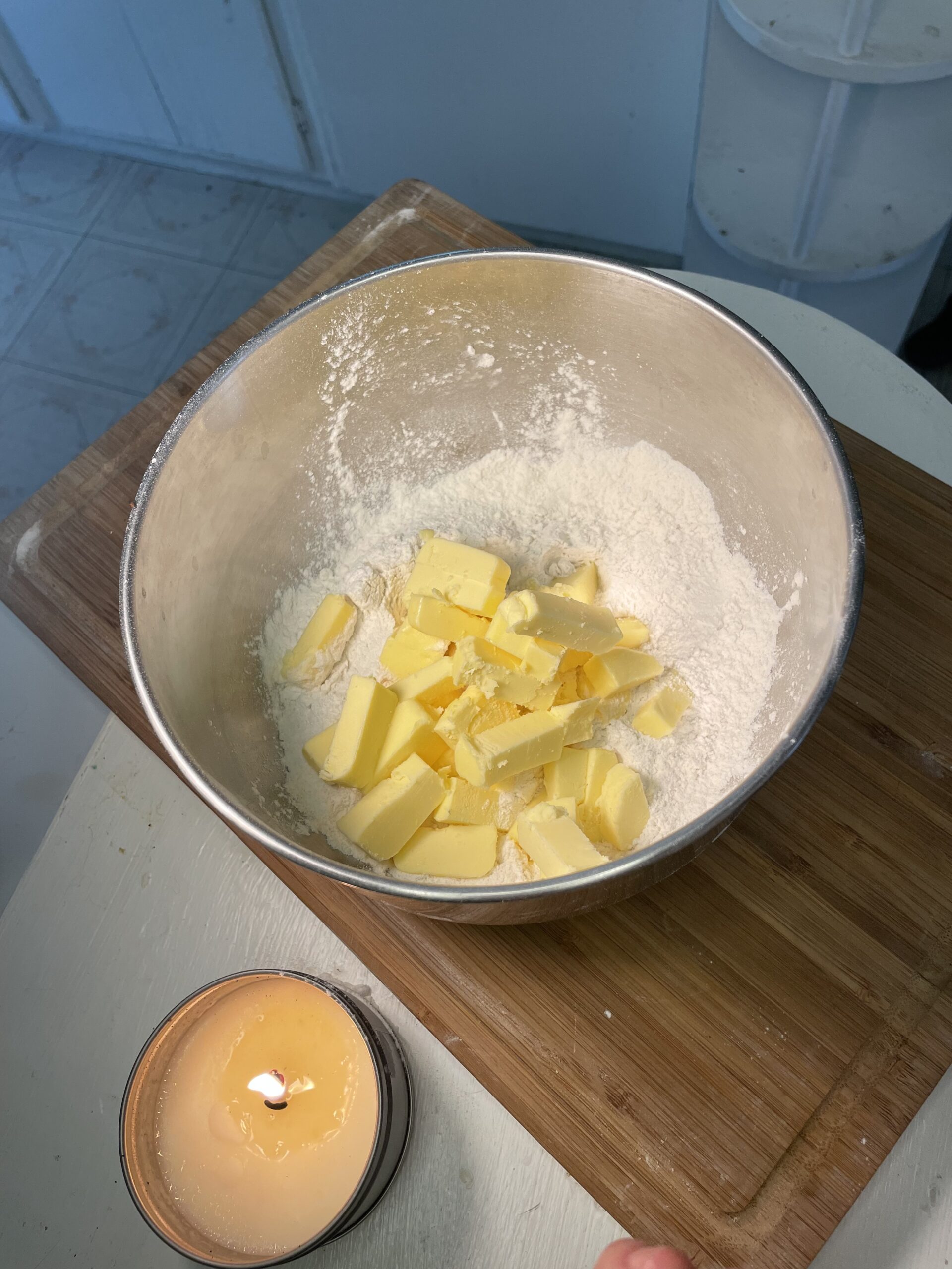 In a light kitchen, there is a wooden cutting board and a lit candle on a white counter. On the cutting board is a large metal mixing bowl with white flour and yellow butter in cubes.