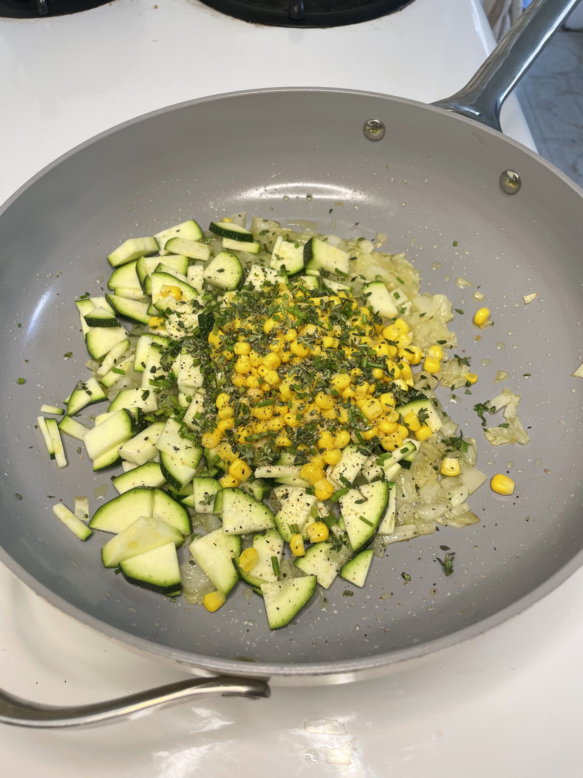 In a light kitchen, a light grey pan is cooking white onion, yellow corn, green zucchini, and herbs. 