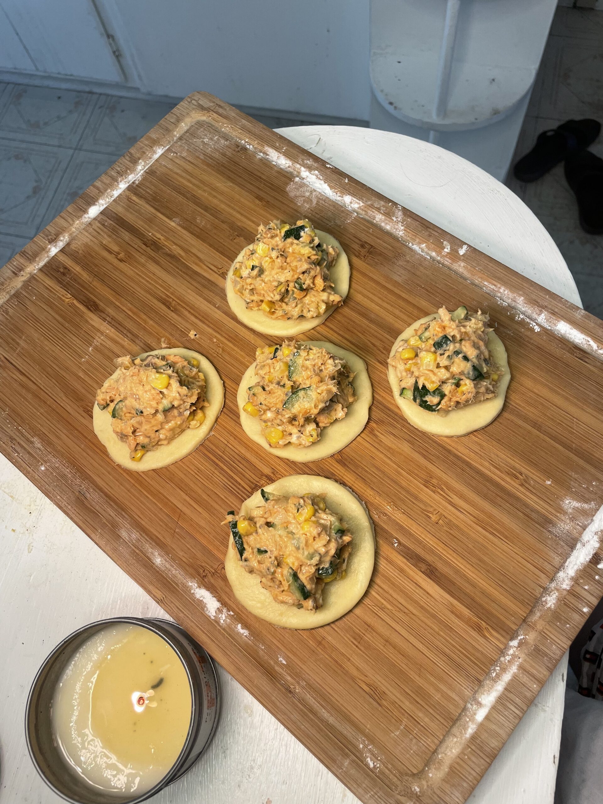 In a light kitchen, there is a wooden cutting board with white flour dusted on it. There are five pie dough rounds with the salmon, veggie mixture on top. You can see pink, orange, green, and yellow throughout the mixture. 