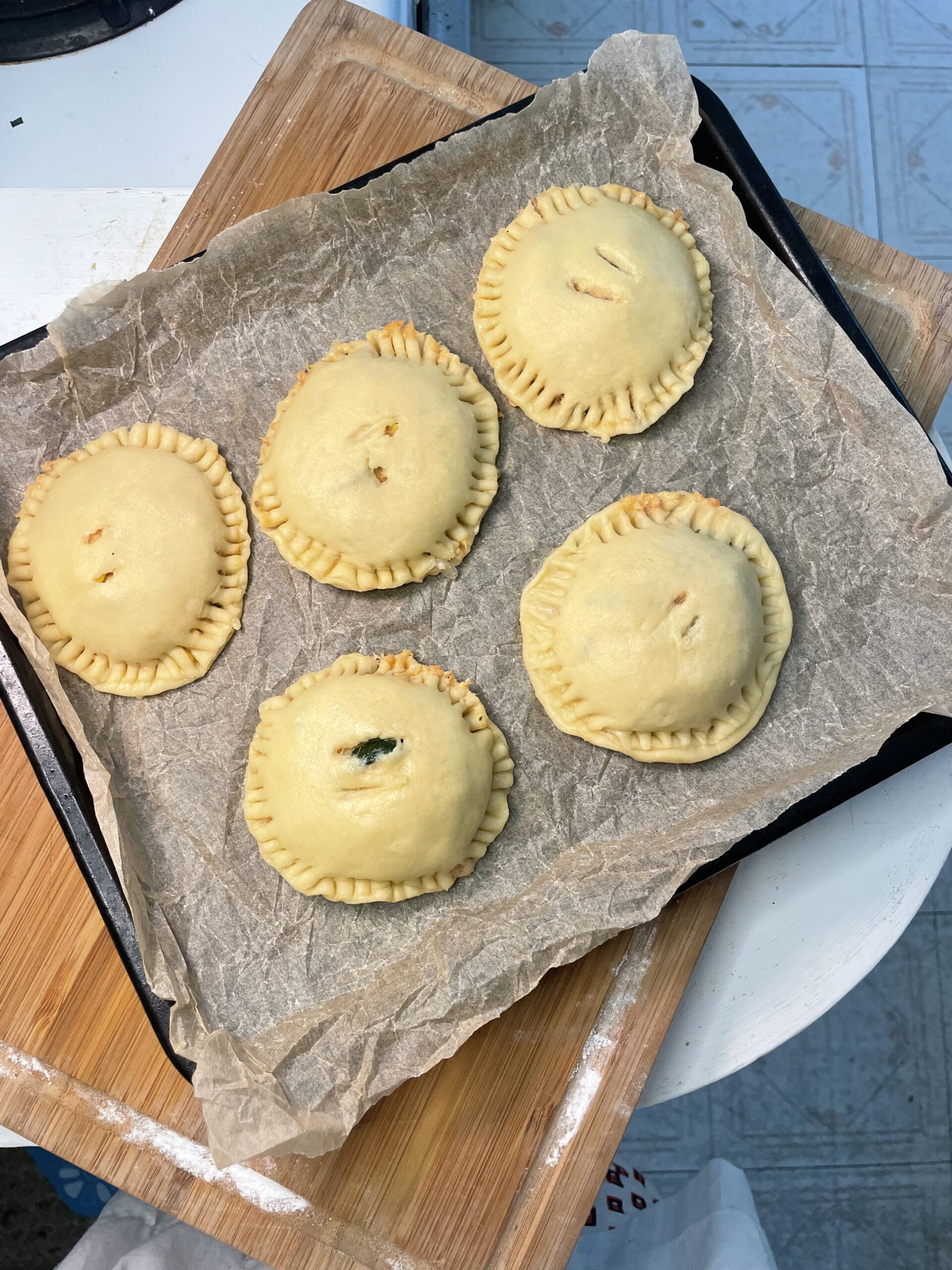 In a light kitchen, there is a wooden cutting board. On top of the cutting board is a black baking sheet with a crumpled parchment paper covering the baking sheet. There are five hand pies on top of the paper. 