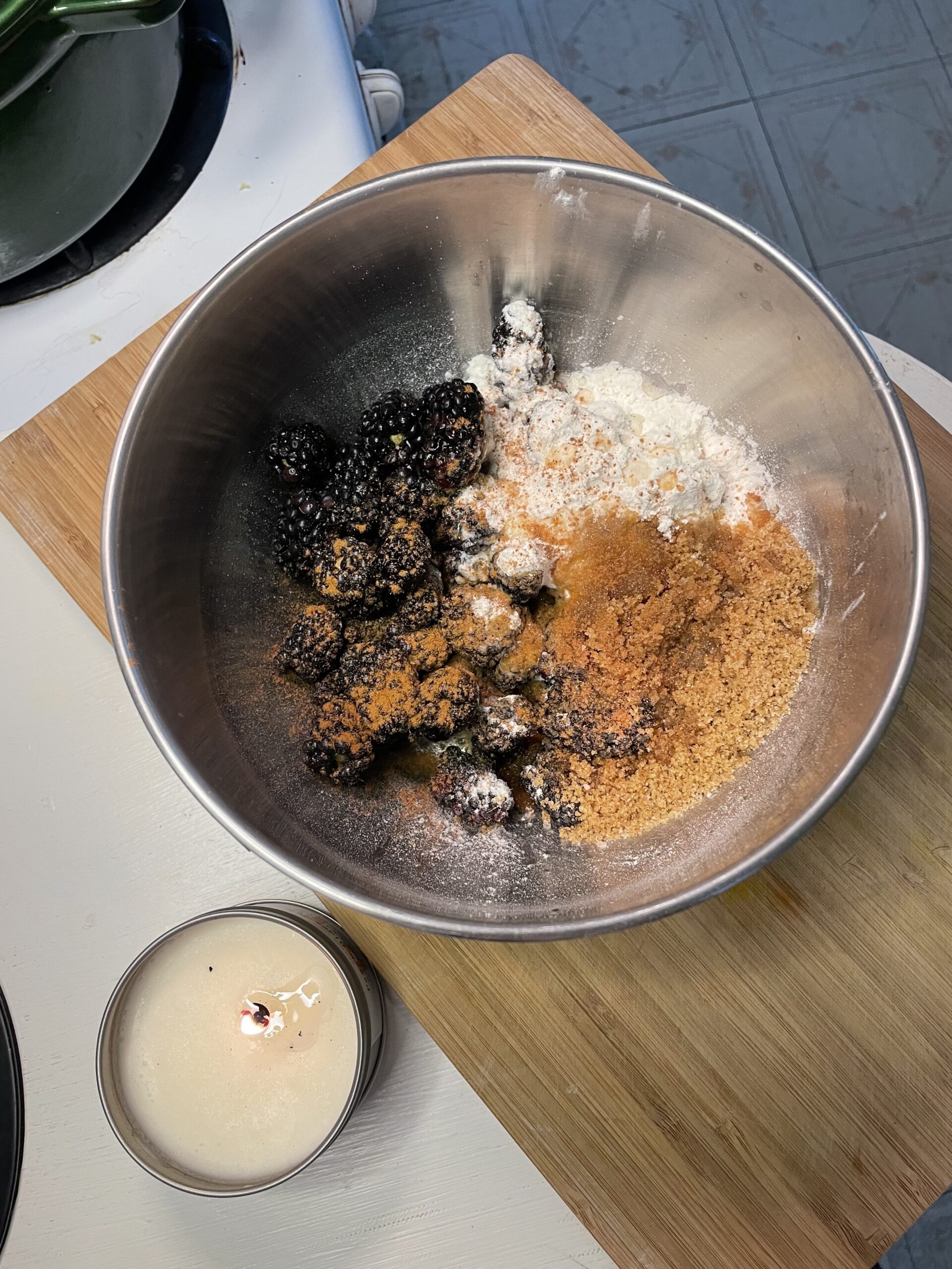 A stainless steel mixing bowl holds fresh blackberries mixed with a combination of dry ingredients, likely including brown sugar, granulated sugar, flour or cornstarch (as a thickener), and warm spices like cinnamon or nutmeg. The mixture is not yet stirred, showing a beautiful contrast between the dark berries and the various powdery ingredients.