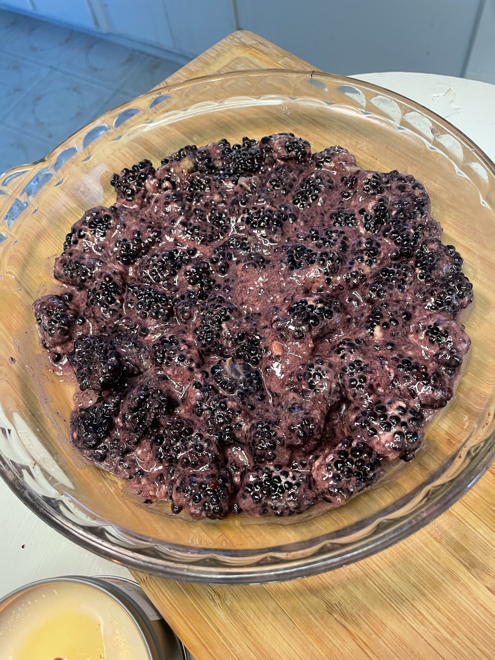 The blackberry mixture has been combined and spread evenly in a clear glass pie dish. The berries now appear juicy and glossy, coated in a thick, sweet filling that’s ready for a topping or baking. You can also see a lit candle in the corner, adding a cozy and homestyle vibe to the preparation setting.