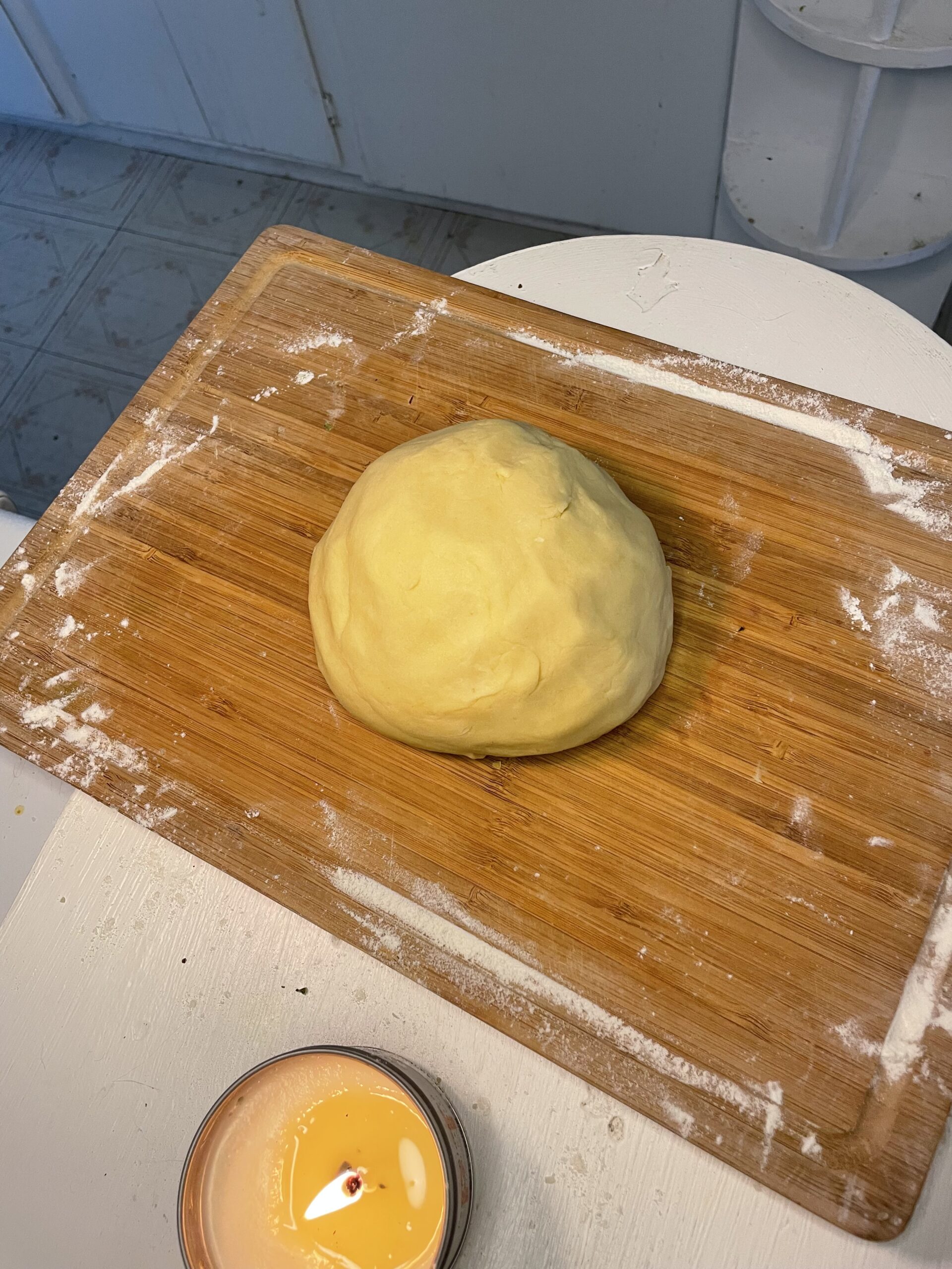 This image captures the dough and crust process for this blackberry cobbler recipe. The dough is in a round ball, placed on a floured, wooden cutting board. 
In the lower left of the image, a lit candle adds a warm, cozy atmosphere, emphasizing the homemade, comforting vibe of the dish.