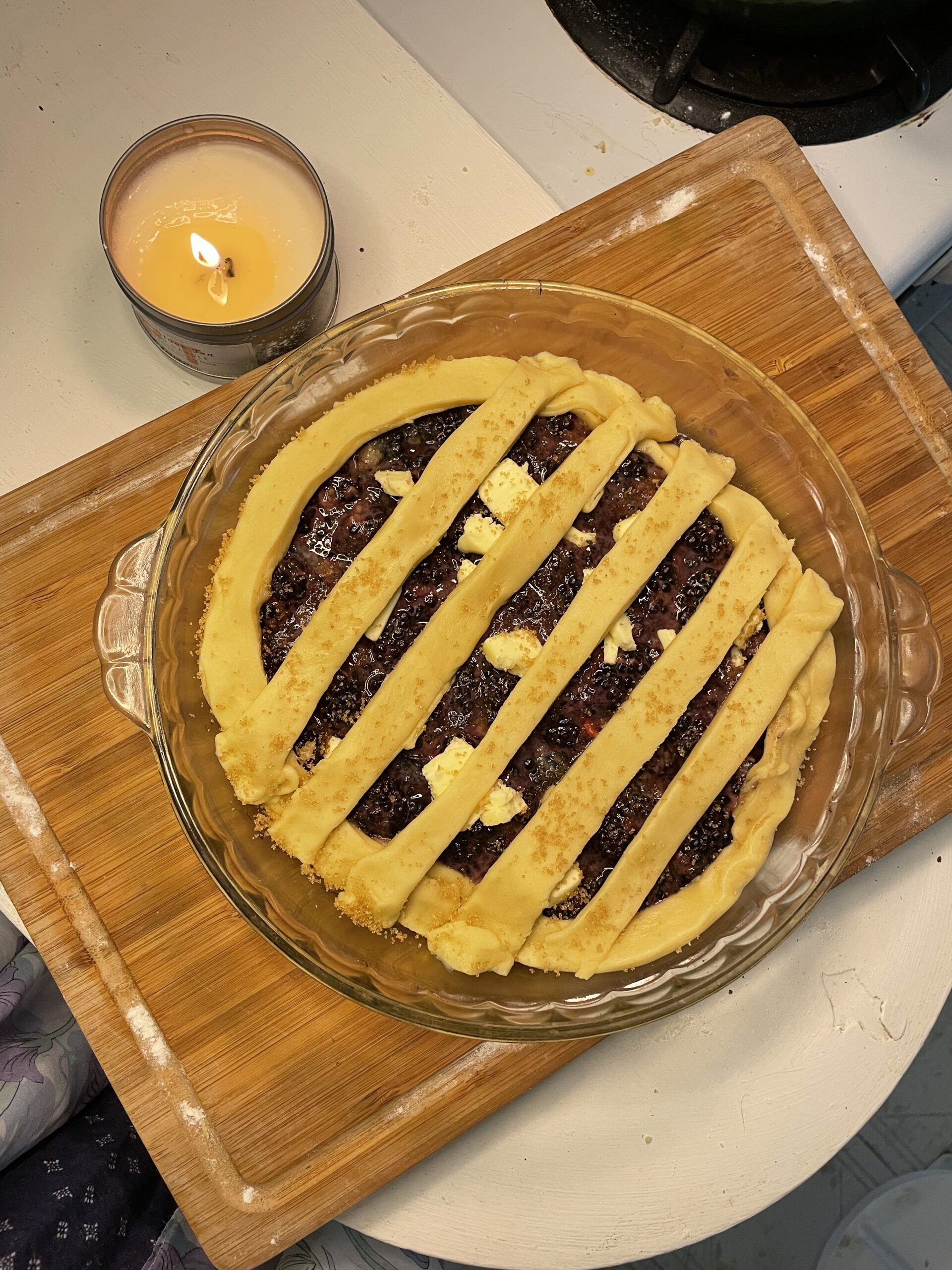 This image captures an assembled, uncooked blackberry cobbler just before baking. The cobbler is in a clear glass pie dish, placed on a wooden cutting board. The rich, dark blackberry filling is visible underneath a decorative top crust.
The crust is laid out in a lattice pattern, using wide, hand-cut dough strips that are slightly rustic and homey. Dabs of butter have been added on top of the berry filling, likely to enhance richness and promote caramelization while baking. The crust appears to be lightly sprinkled with sugar, which will likely create a golden, crispy finish once baked. In the upper left of the image, a lit candle adds a warm, cozy atmosphere, emphasizing the homemade, comforting vibe of the dish.
