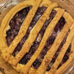 This image captures a fully baked Stardew Blackberry Cobbler just out of the oven. The cobbler is in a clear glass pie dish, placed on a wooden cutting board. The rich, dark blackberry filling is visible underneath a decorative top crust. The crust is laid out in a lattice pattern, using wide, hand-cut dough strips that are slightly rustic and homey. The crust is golden brown and crispy. In the upper left of the image, a lit candle adds a warm, cozy atmosphere, emphasizing the homemade, comforting vibe of the dish.