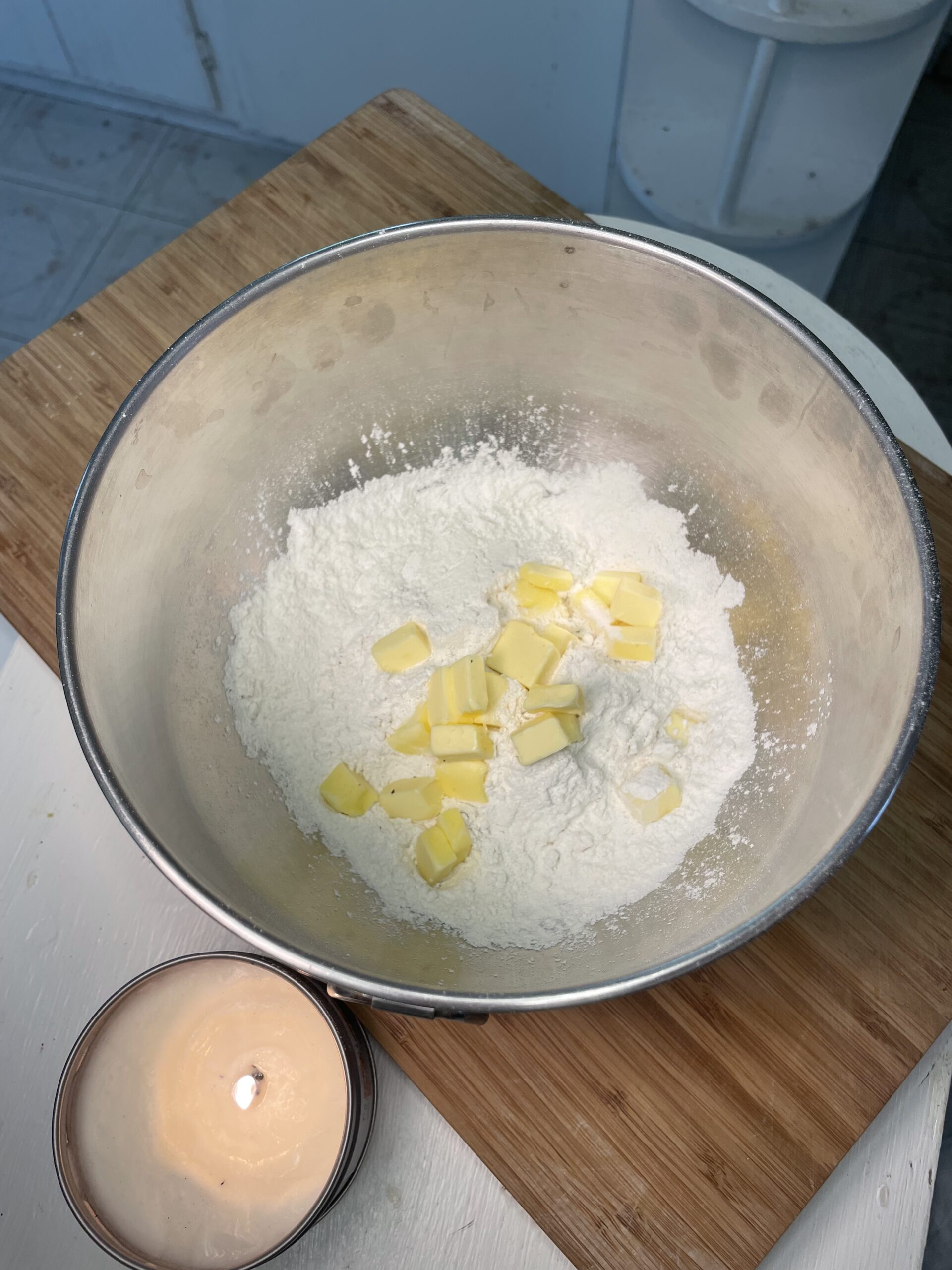 In a light kitchen, there is a wooden cutting board and a lit candle on a white counter. On the cutting board is a large metal mixing bowl with white flour and yellow butter in cubes.