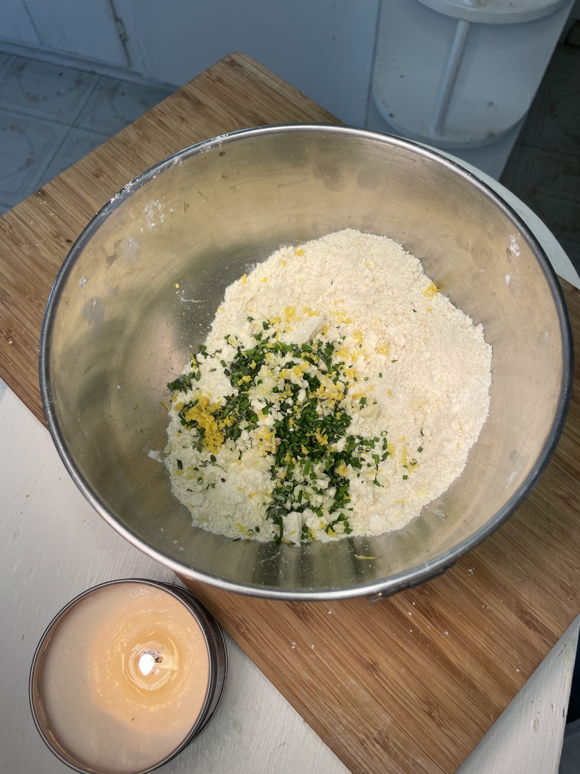 In a light kitchen, there is a wooden cutting board and a lit candle on a white counter. On the cutting board is a large metal mixing bowl with white flour and yellow butter mixed together and herbs added to the mixture.