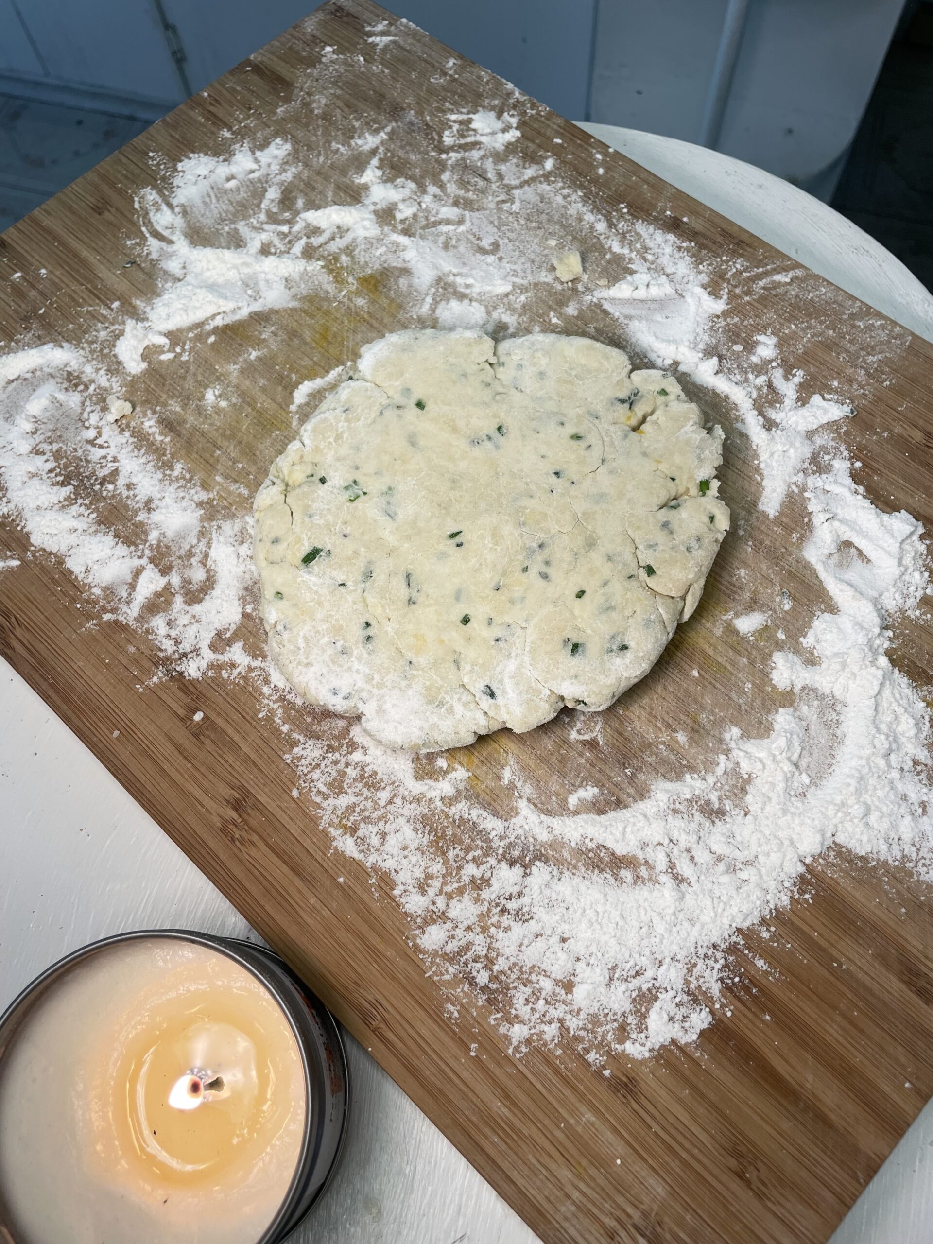 On a white countertop, there is a lit candle and a wooden cutting board. On the cutting board there is flour covering the surface and biscuit dough speckled with herbs rolled out.