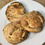 Three expertly baked golden-brown biscuits are stacked on top of each other on a white plate on a wooden cutting board next to a lit candle on a white countertop